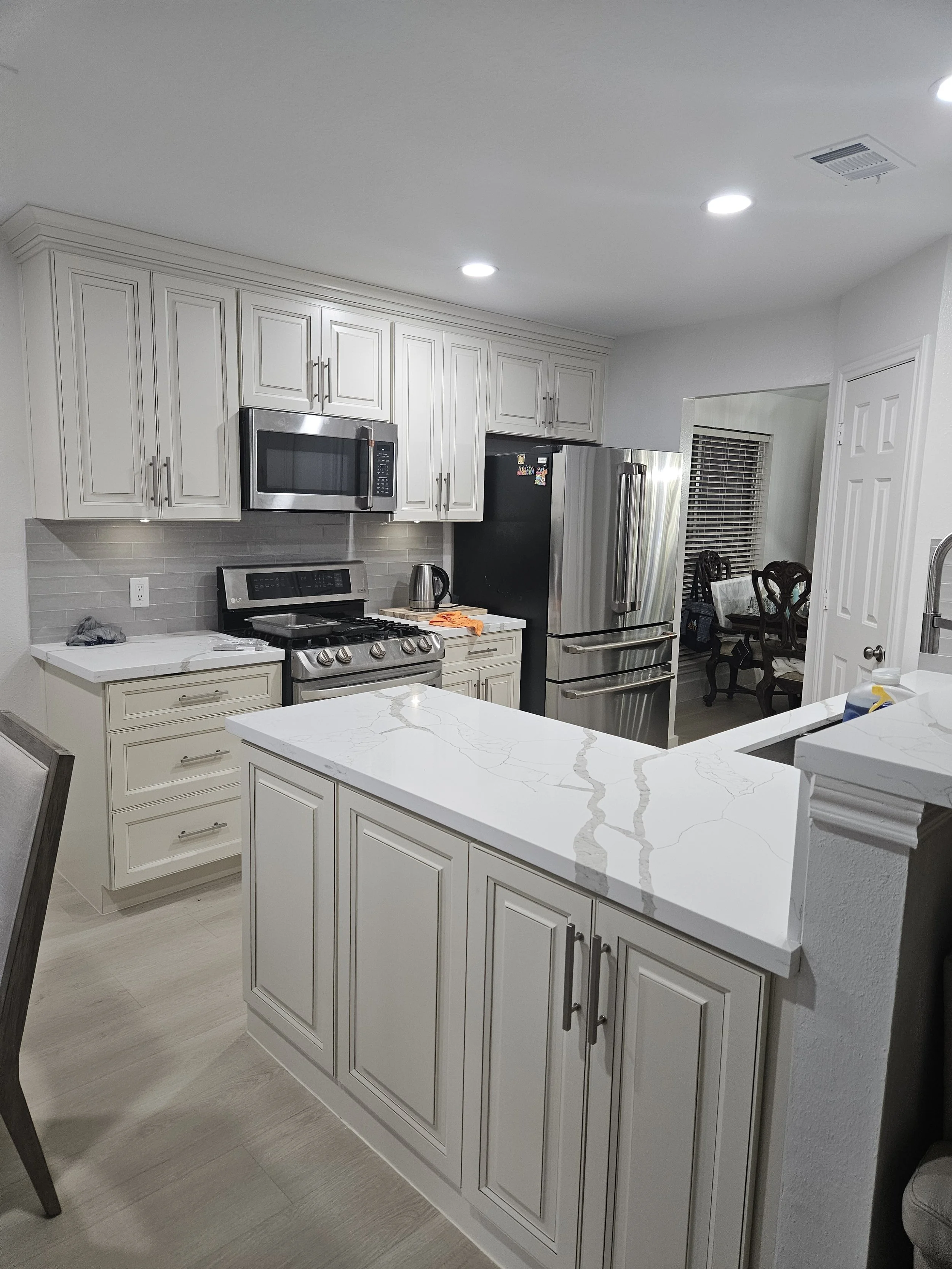 Kitchen remodeling with white cabinets, stainless steel refrigerator, microwave, stove, and marble countertop. Dining table with chairs, window with blinds, and ceiling lights.