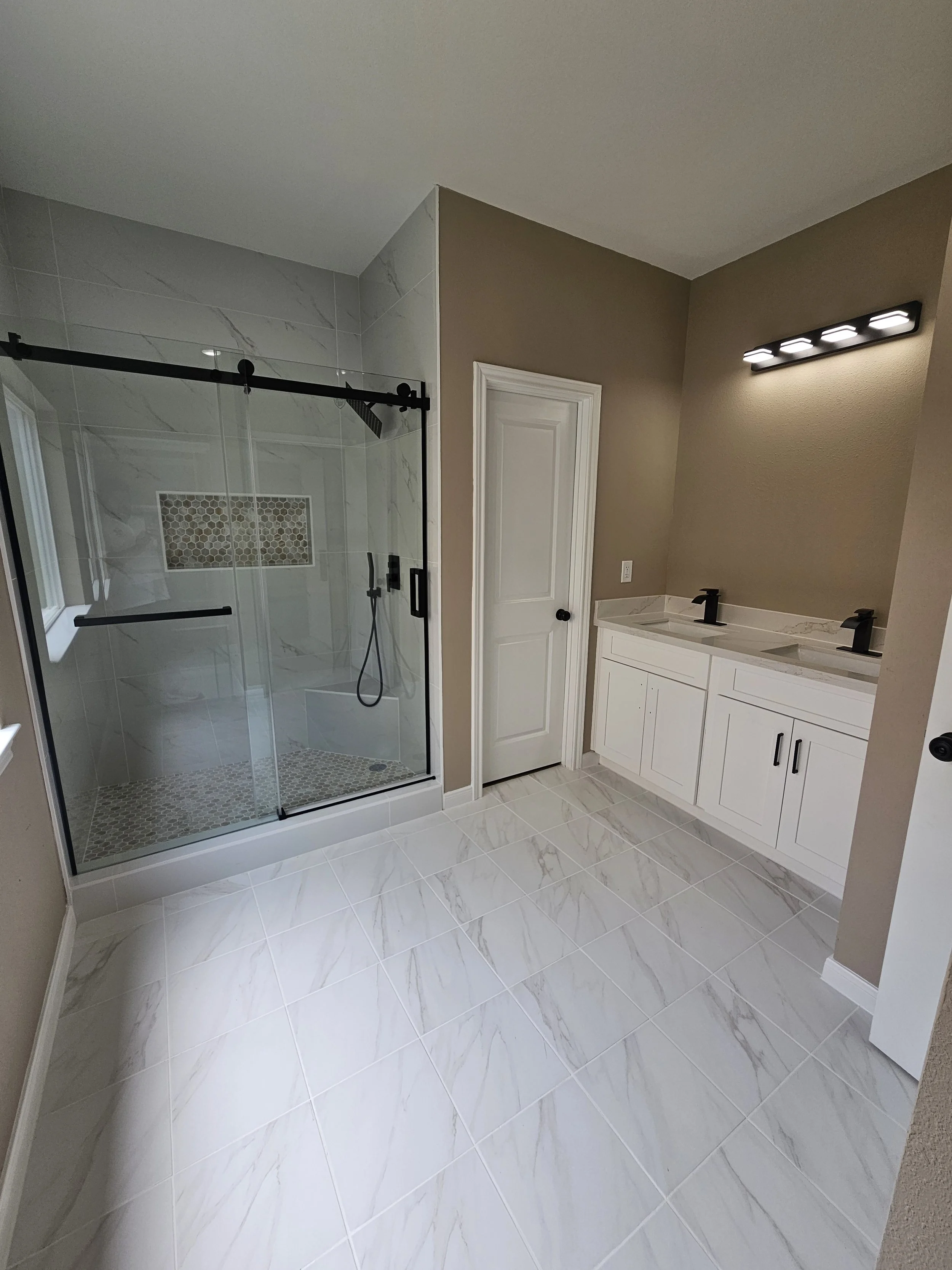 Modern bathroom with a glass-enclosed shower with black hardware, a double vanity with black fixtures, white cabinetry, marble countertops, and beige walls.