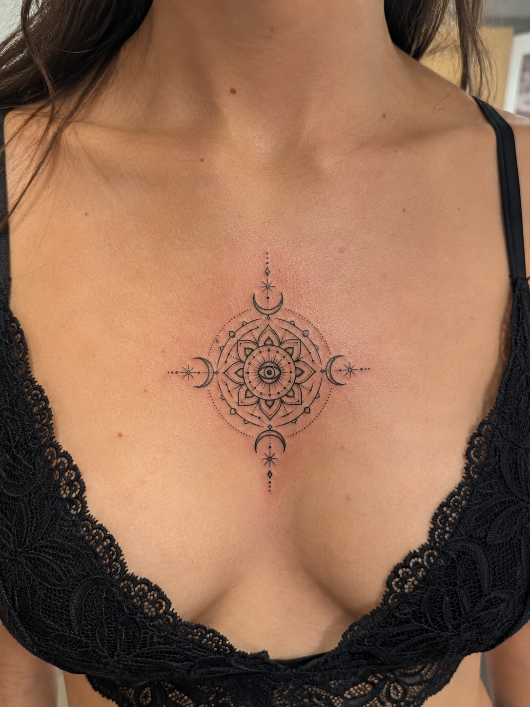 Tatouage de style géométrique et mystique sur la poitrine d'une femme, comprenant une lune, des étoiles, un œil au centre, et des motifs symétriques.