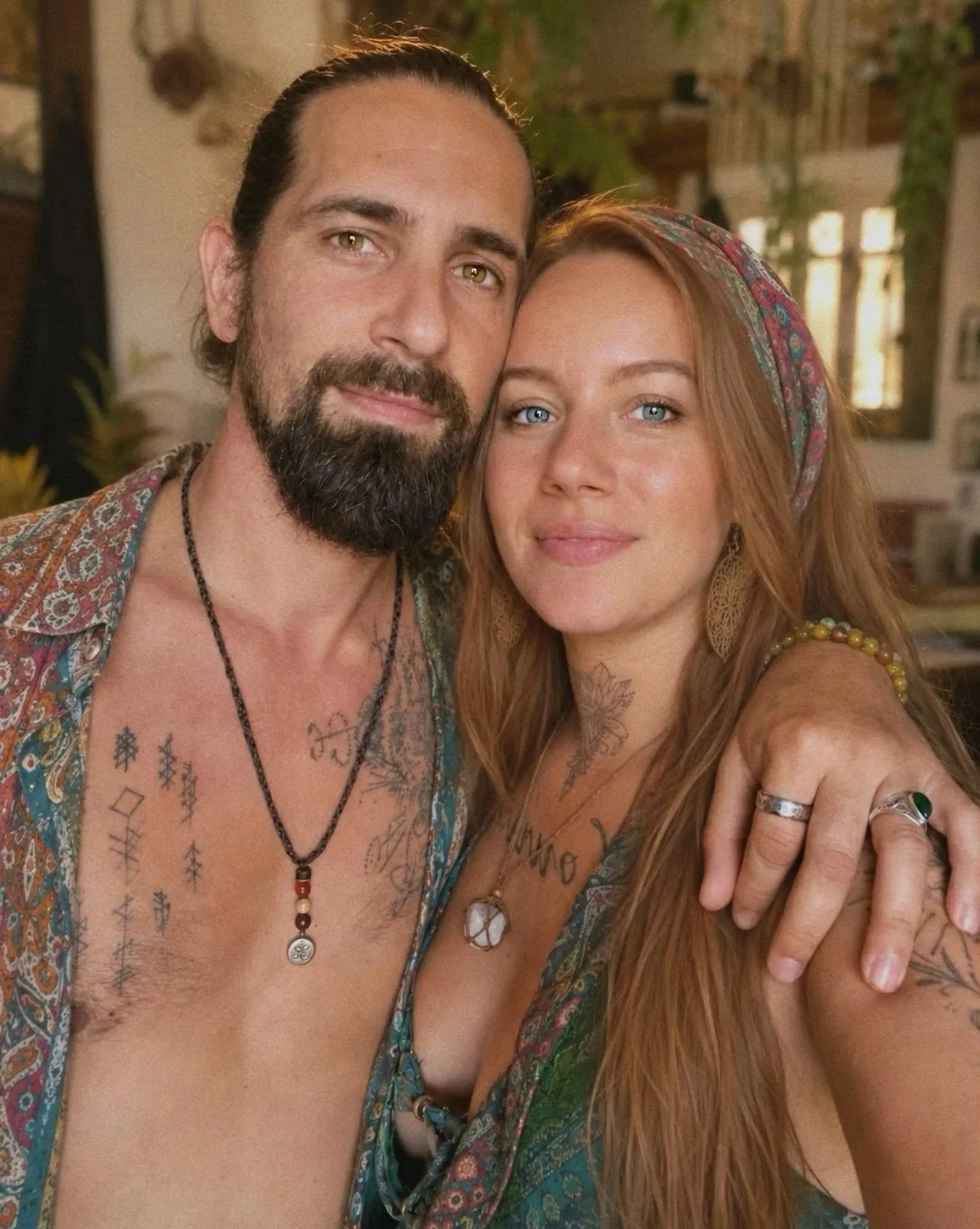 Un homme avec une barbe et une femme aux cheveux roux prennent une selfie ensemble à l'intérieur, souriants et proches, avec des bracelets, des colliers et des tatouages visibles.