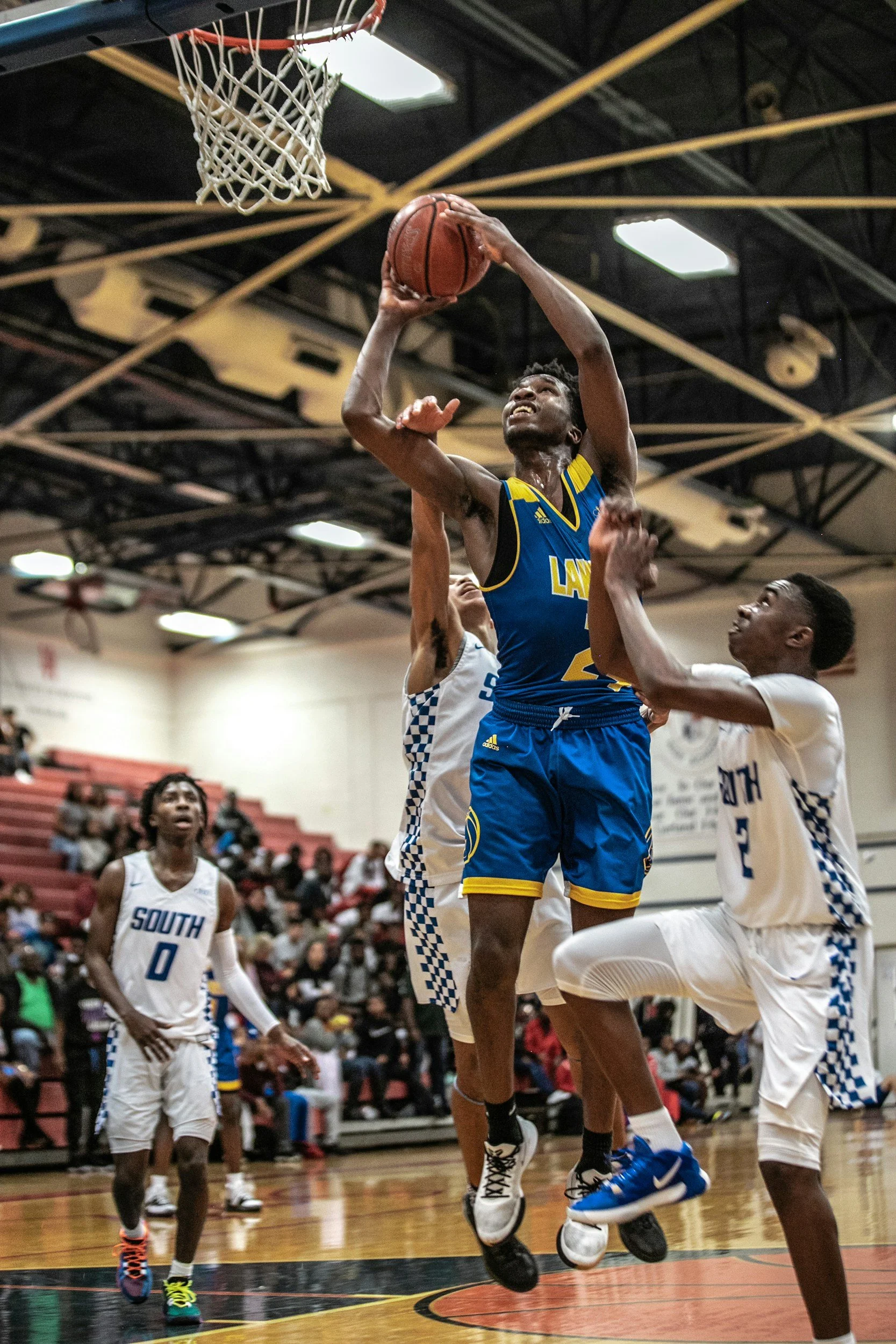 A basketball player in a blue jersey is jumping to make a shot while two defenders in white jerseys try to block him during a game. The game is taking place in a gymnasium with an audience in the background.