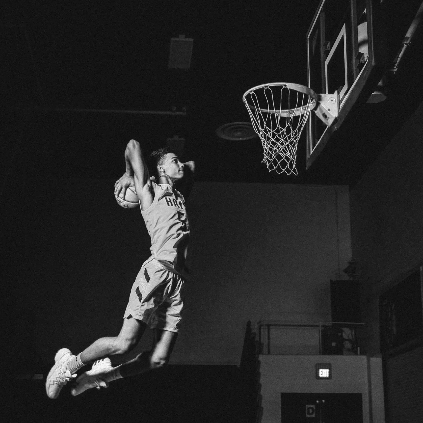 Black and white photo of a basketball player jumping towards the hoop for a dunk shot in an indoor gym.