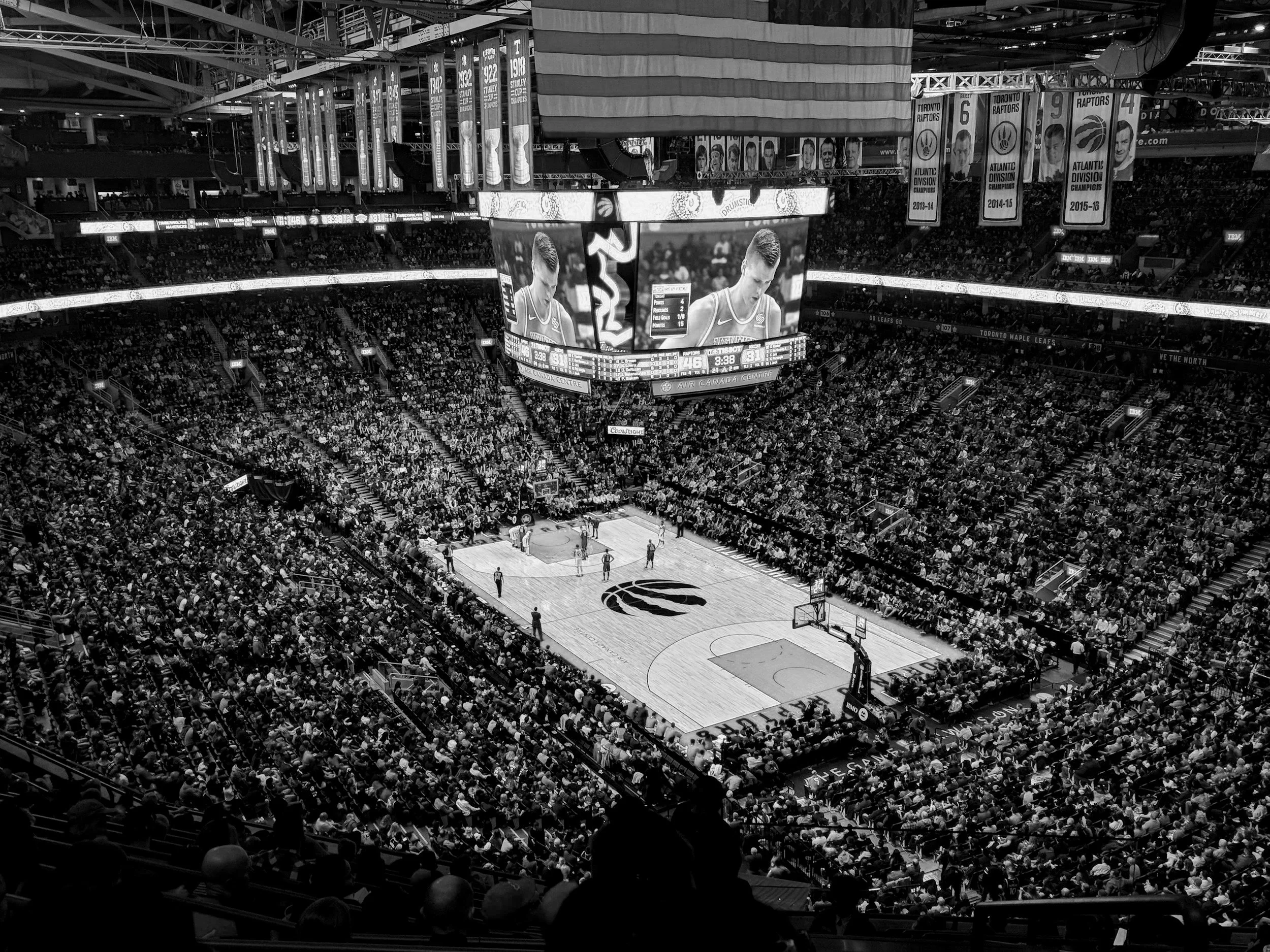 Inside a large basketball arena filled with spectators watching a game. The court features the Toronto Raptors logo, and there is a large screen showing a player in a Raptors jersey. Banners hang from the ceiling celebrating the team's championships.