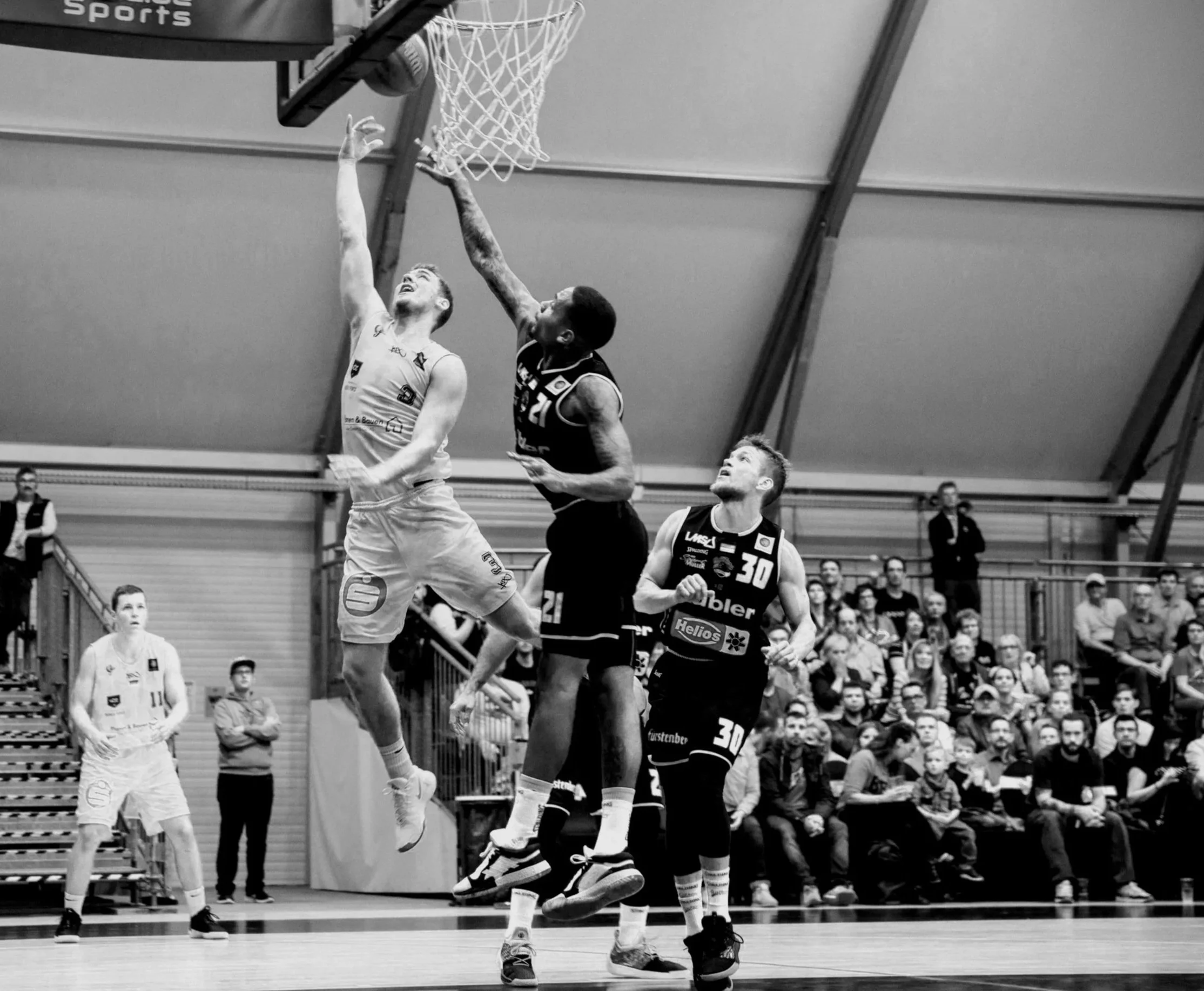 Two basketball players with athletic builds jumping to reach the ball near the hoop, while other players and spectators observe in the background.