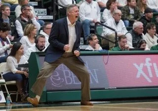 Steve Barnes in a suit is shouting and holding onto the court railing during a basketball game, with an audience in the stands watching.