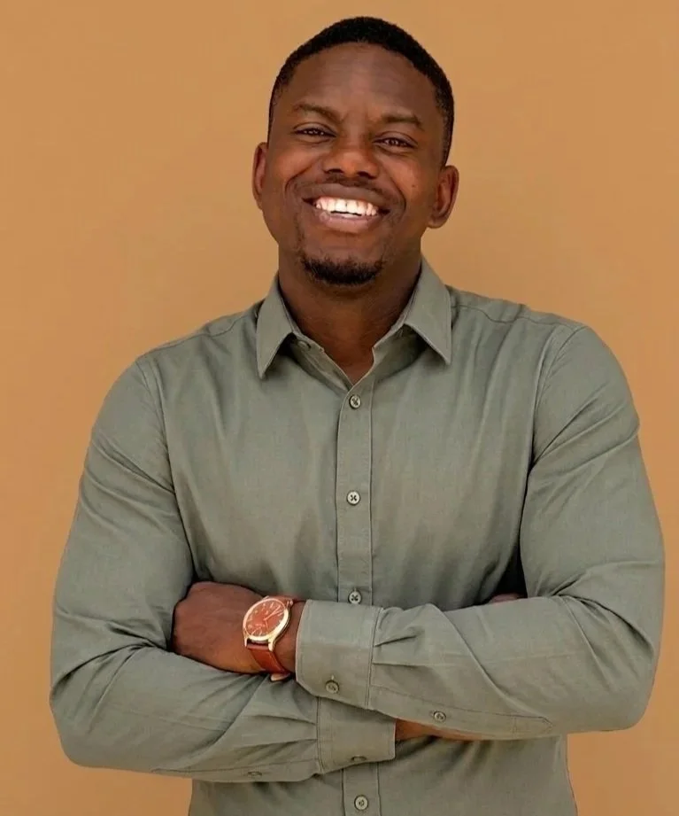A smiling man with dark hair and a beard wearing a light green button-up shirt and a brown watch, standing with arms crossed against a beige background.
