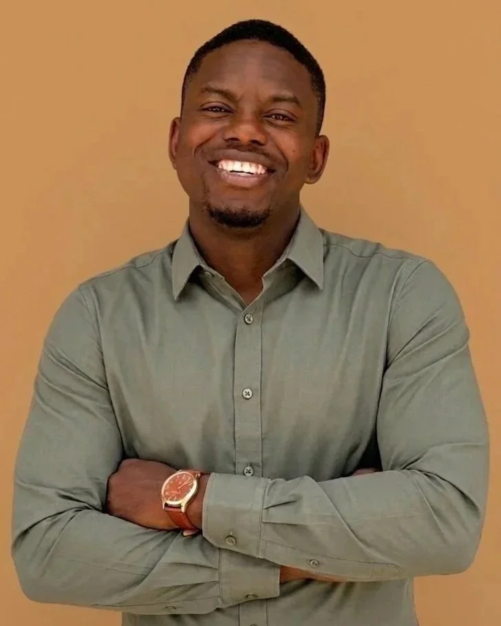 A smiling man with crossed arms, wearing a gray button-up shirt and a watch, standing against a beige background.