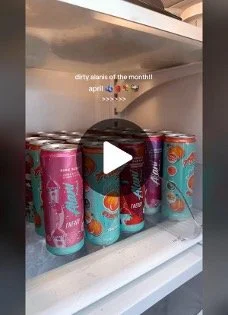 A refrigerator shelf holding multiple cans with colorful branding, some with pink and blue designs. Text overlay says, 'dirty alias of the month lol april' with sunset and fire emojis.