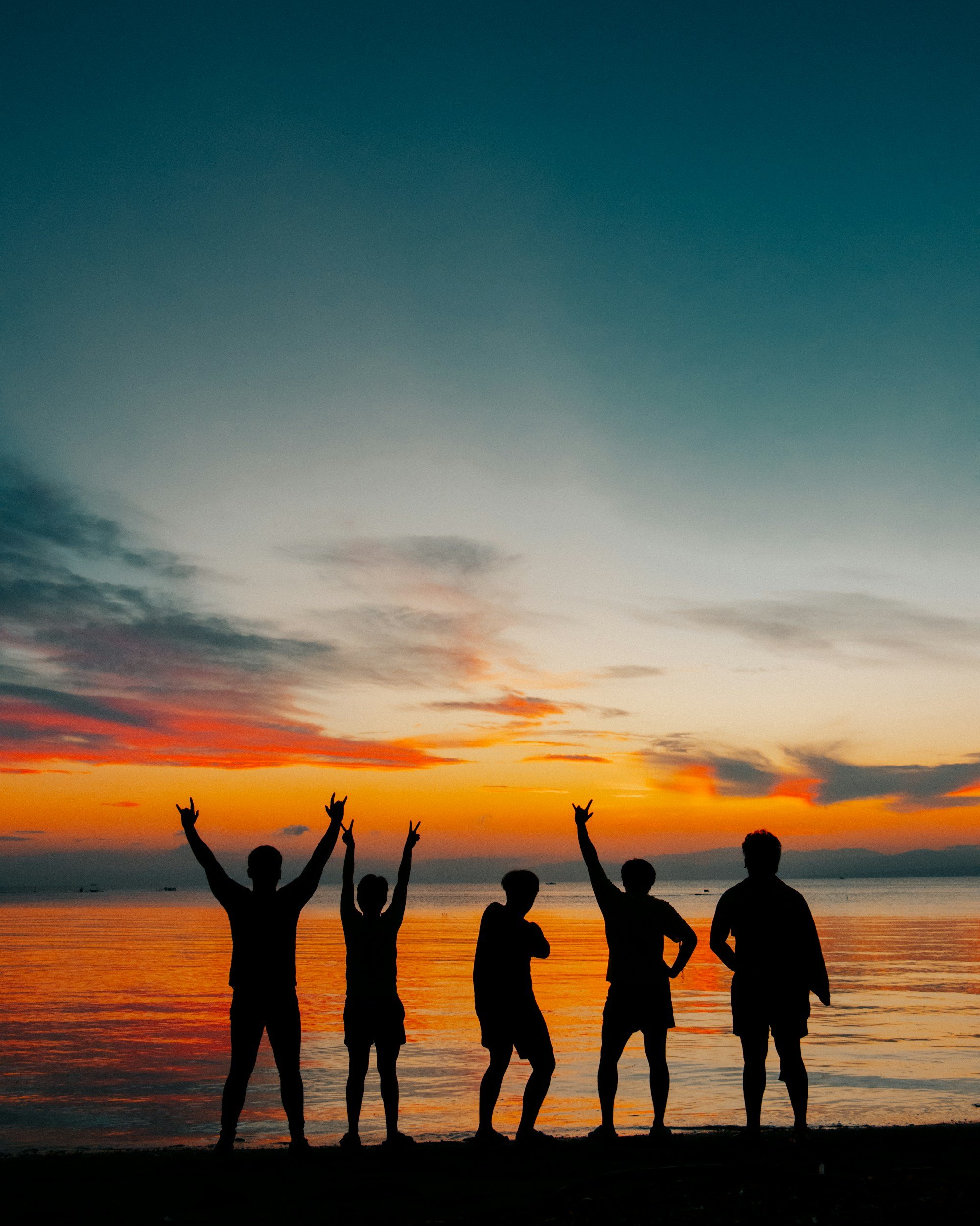 People silhouetted against a colorful sunset over the ocean, making peace signs and striking poses on the beach.