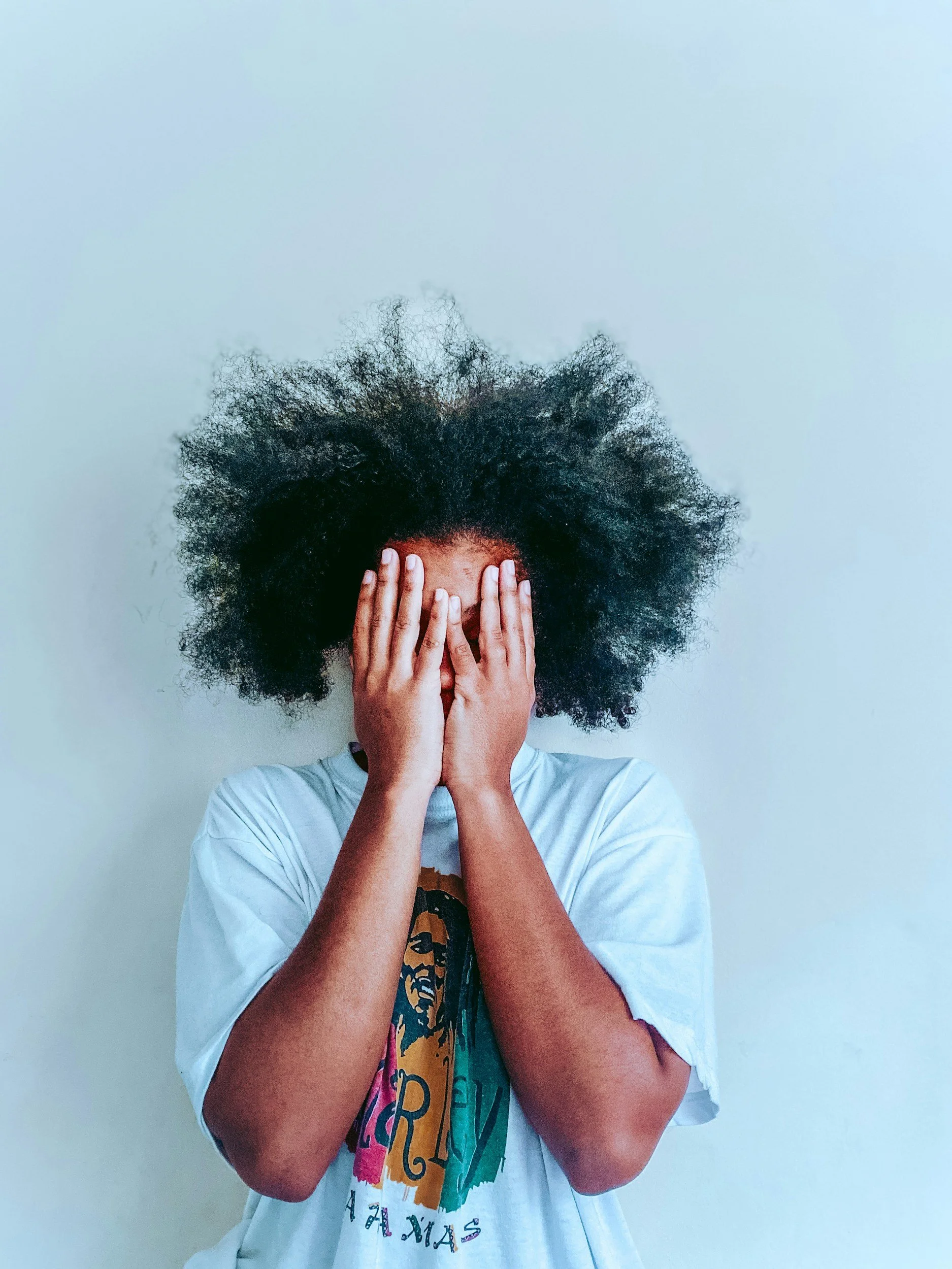A person with curly black hair covering their face with hands, wearing a white T-shirt with colorful text against a plain light gray background.