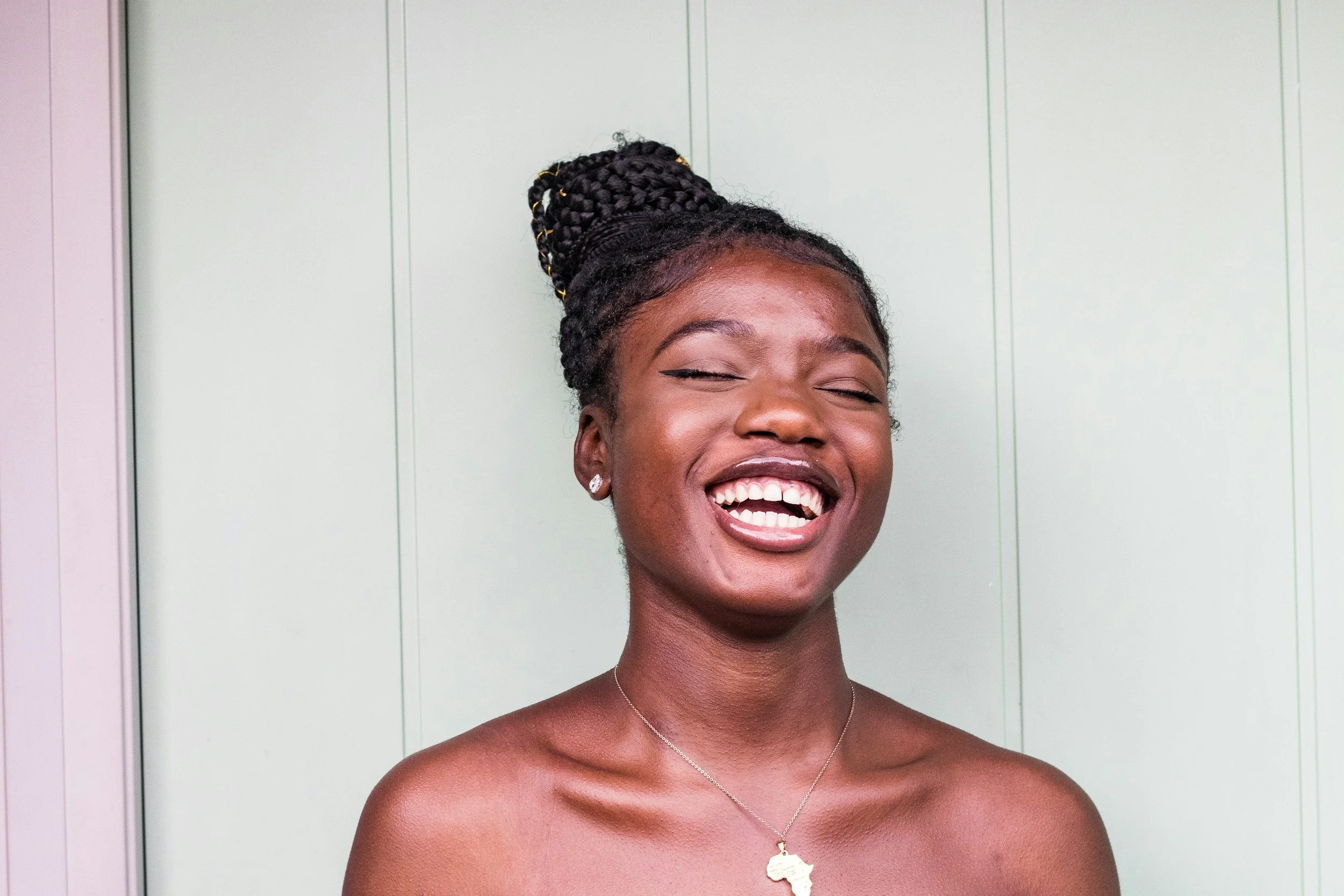 A woman with dark skin, braided hair styled in a high bun with gold accents, smiling widely with eyes closed, wearing earrings and a necklace with an African continent pendant, against a light-colored background.
