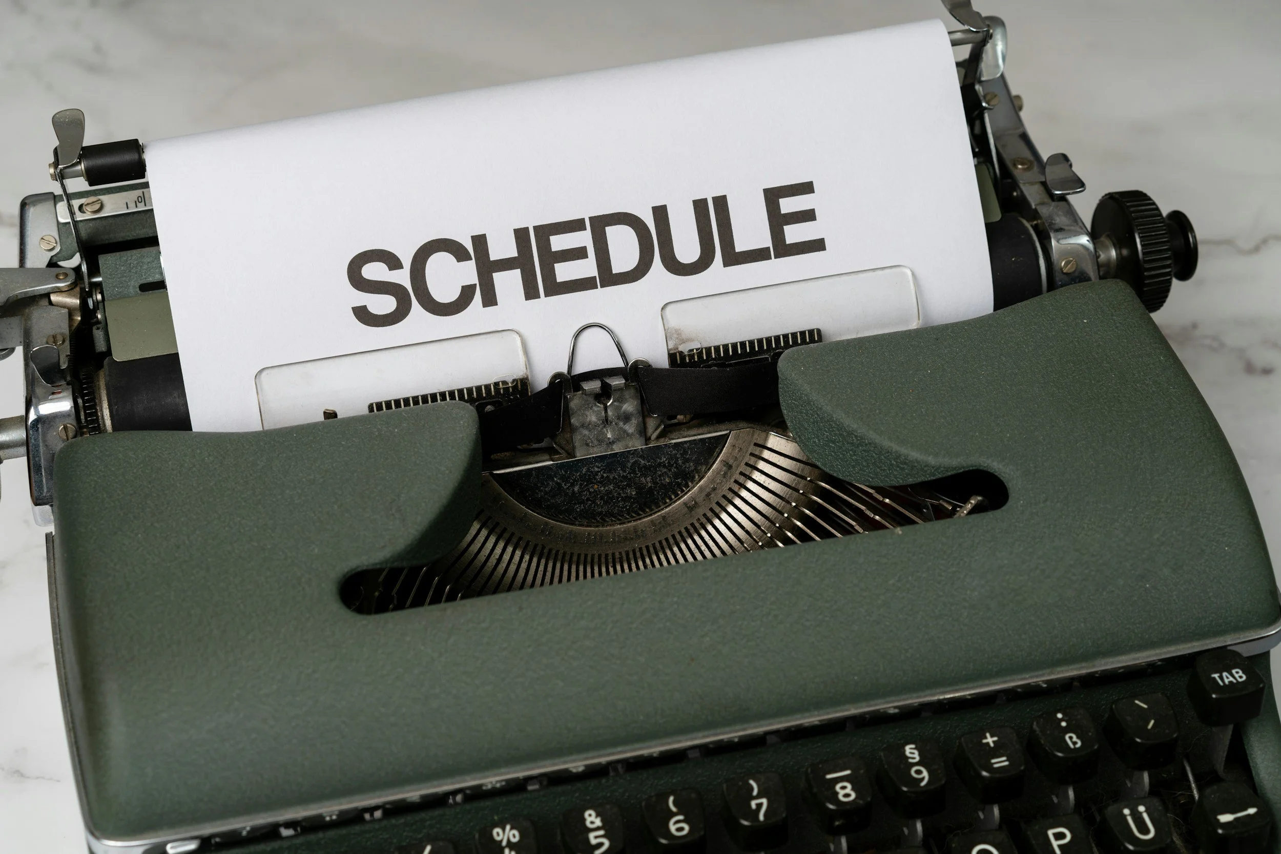 A woman with short blonde hair wearing a sleeveless orange top and beige pants, sitting on a bench, writing in a notebook. There is a large leafy plant on her left and a wooden panel behind her. A shadow of window panes is cast on the plain white wall to her right.
