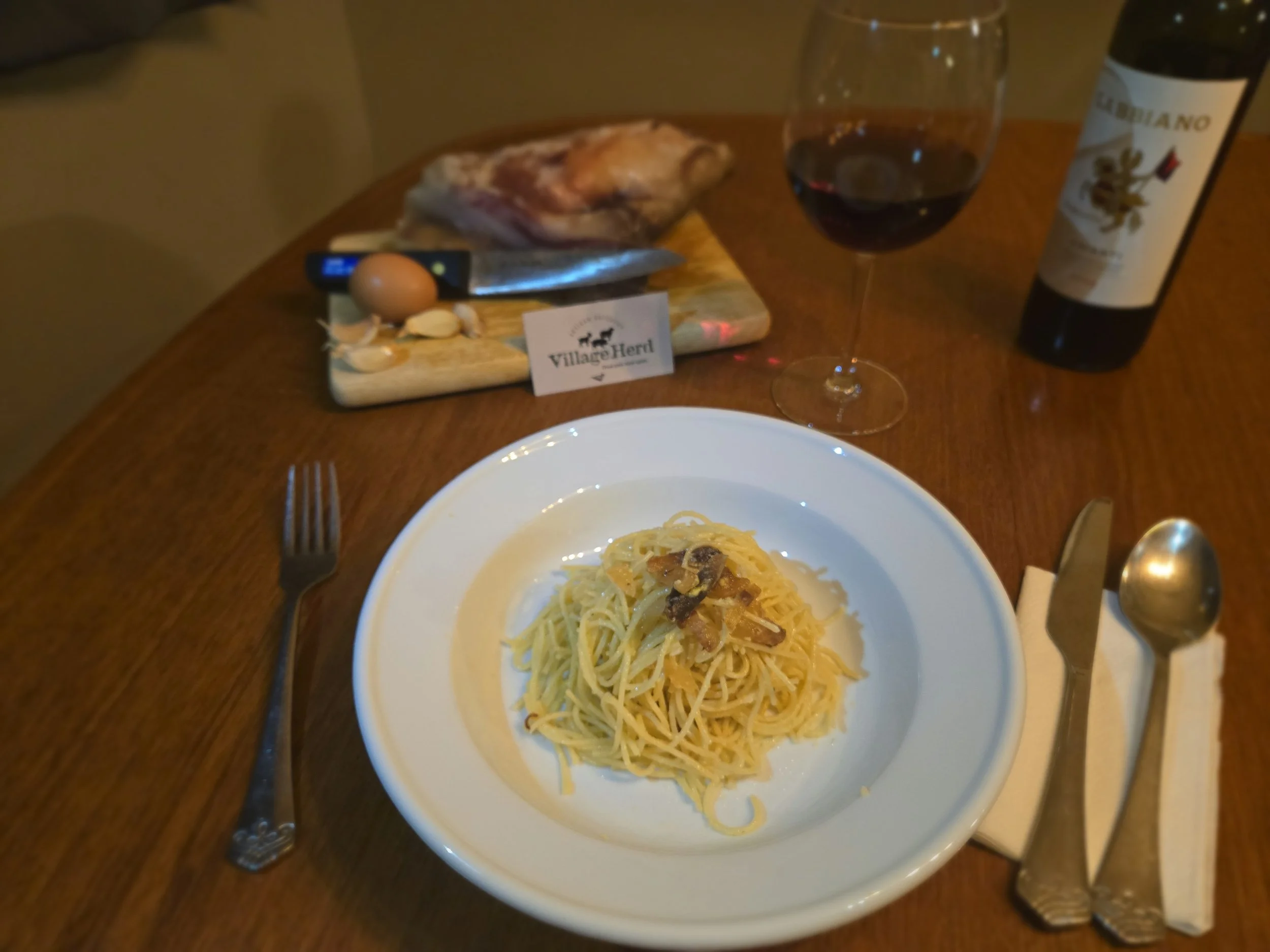 A bowl of pasta carbonara with a glass of wine, ready for dinner. In the background, the guanciale used to make it sits on a cutting board with a brown egg and some garlic cloves.