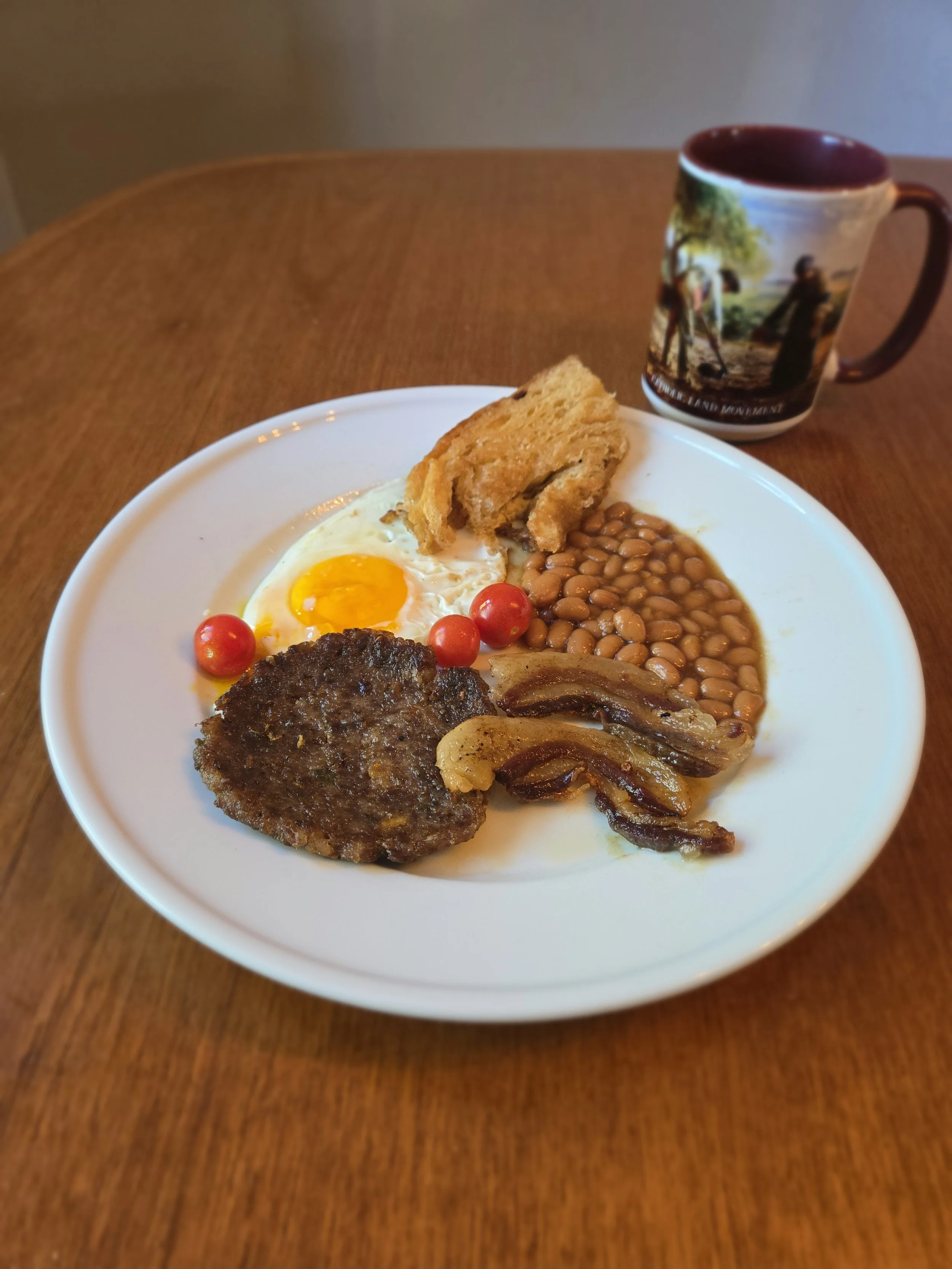 Breakfast sausage patty, bacon, baked beans, sunny-side up egg, and tomatoes on a plate with a coffee mug nearby