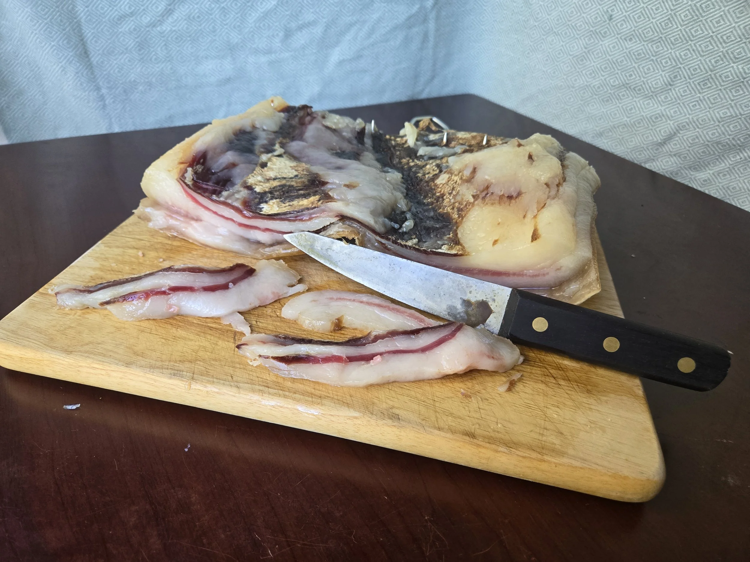 Slab of salt-cured bacon on a cutting board with a few slices removed and a masahiro boning knife