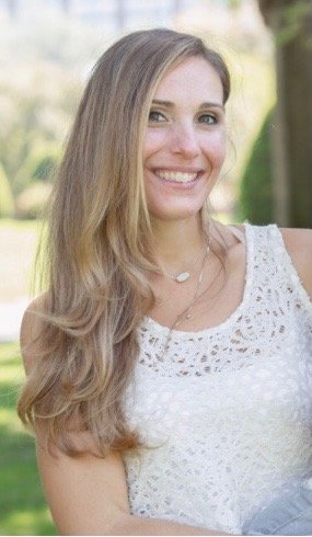 A young woman outdoors with long, wavy light brown hair, smiling, wearing a white lacy top and a necklace, with blurred greenery in the background.