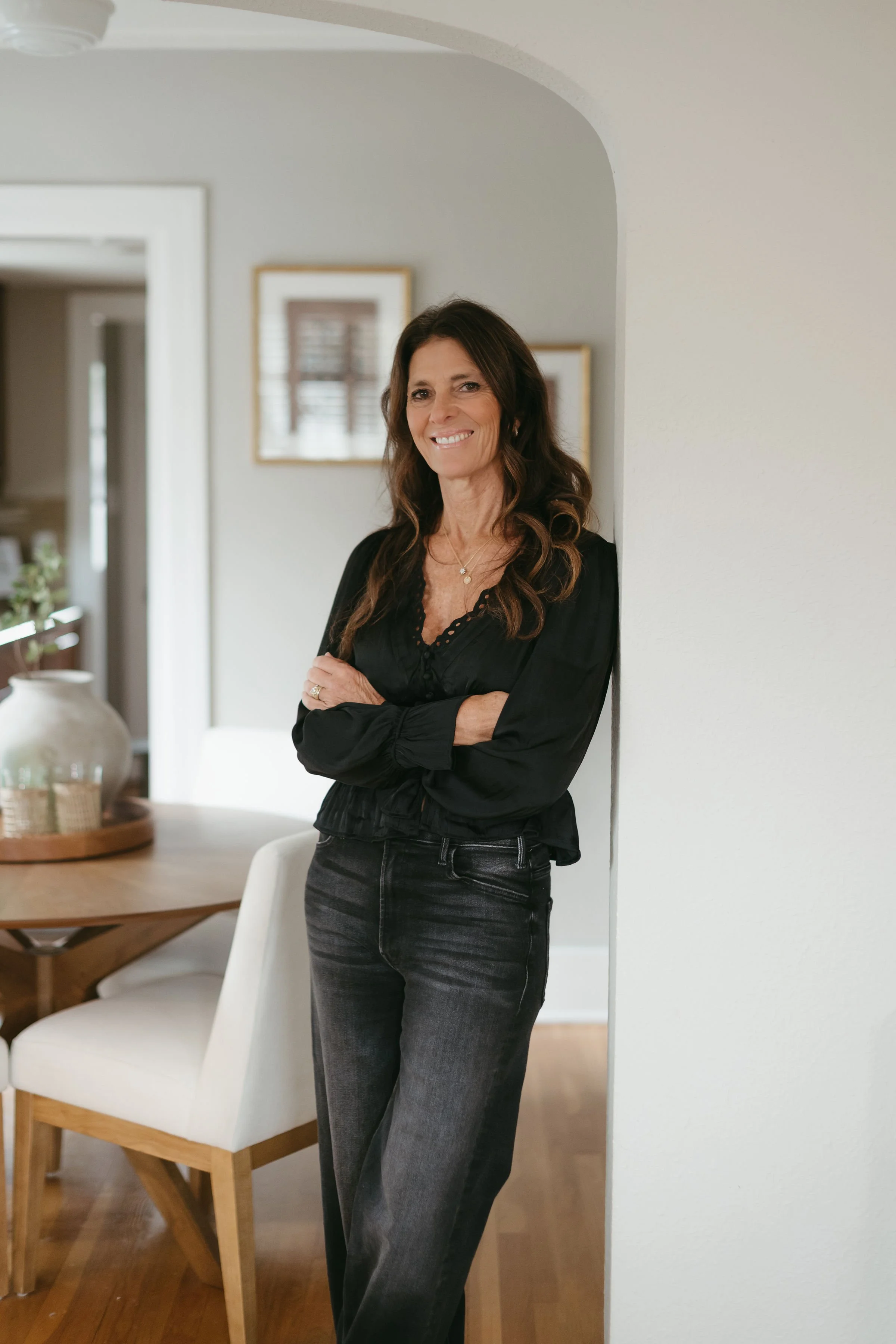 A woman with long brown hair smiling, wearing a black top and dark jeans, standing indoors next to a white wall, with a dining table and chairs visible in the background.