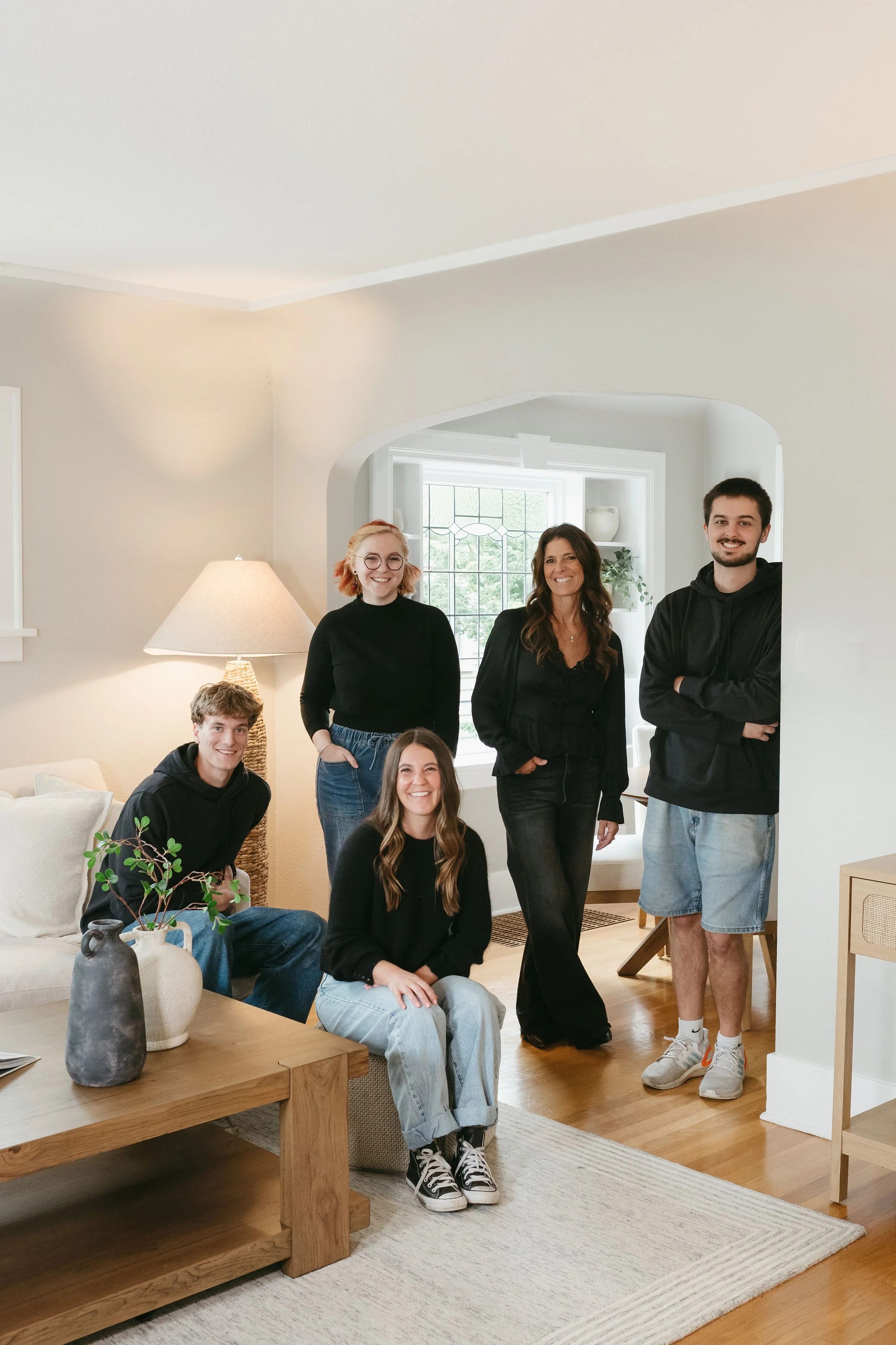 Group of six people, three women and three men, smiling and posing inside a well-lit living room with light-colored walls, wooden flooring, a window with a grid pattern, and a beige area rug. One person is seated on a sofa, and the others are standing.