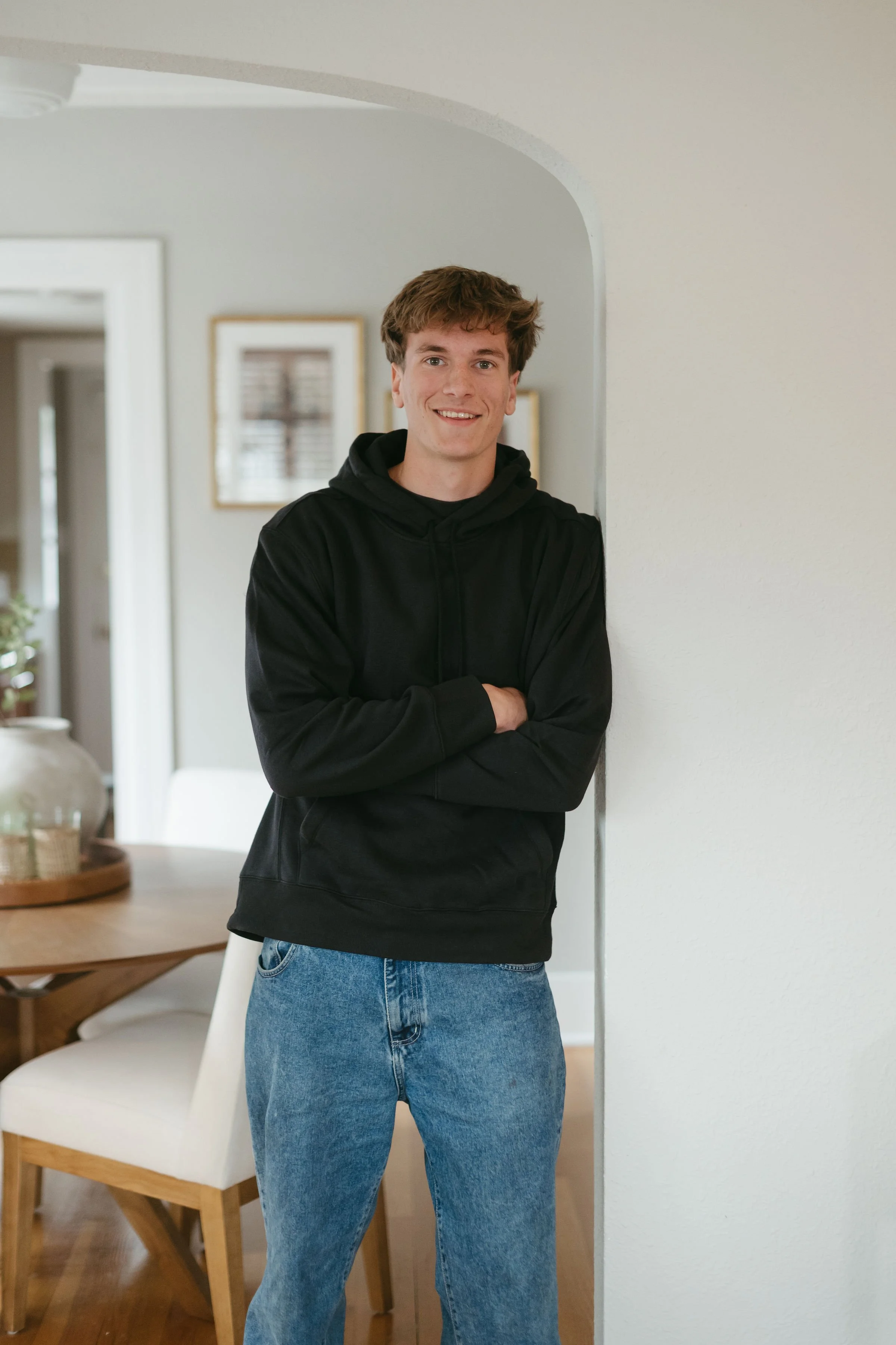 A young man with short brown hair and a light smile, wearing a black hoodie and blue jeans, standing with arms crossed in a well-lit room with white walls and framed pictures in the background.