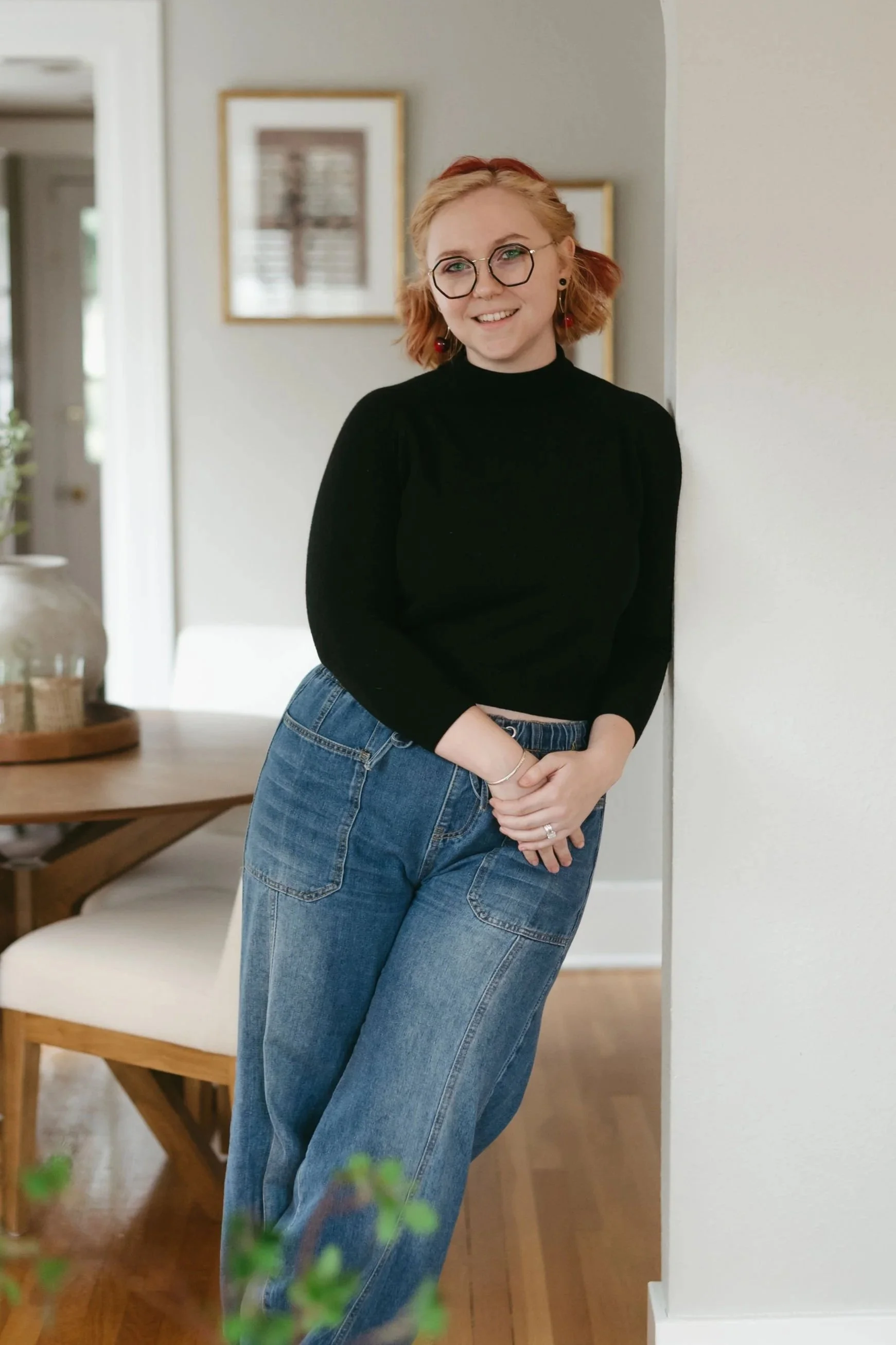 A woman with red hair, glasses, and earrings smiling and leaning against a wall in a living room with a wooden table and framed pictures in the background.