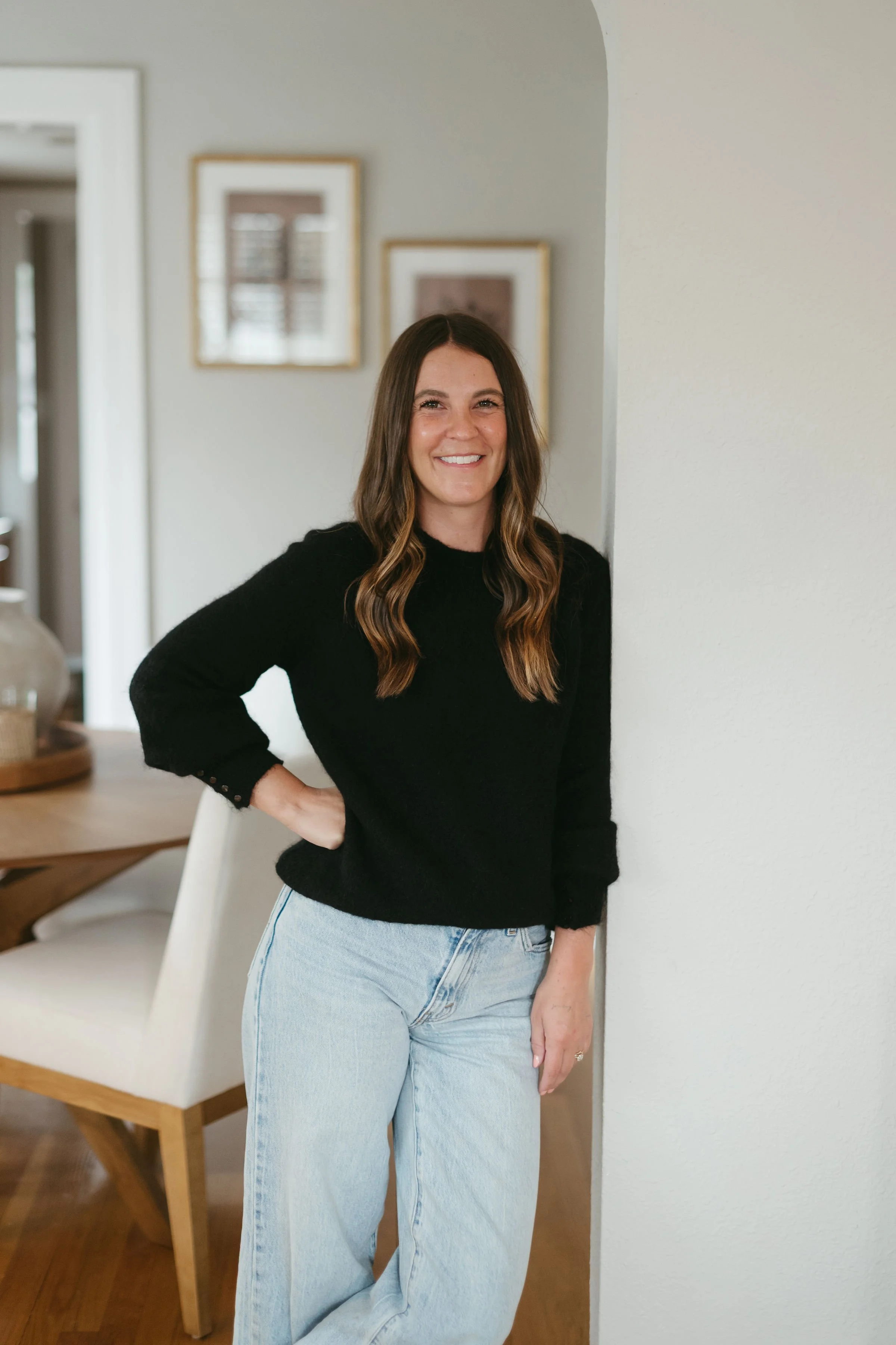A woman with long wavy brown hair wearing a black sweater and light blue jeans standing in a room with a table and chairs, smiling at the camera.