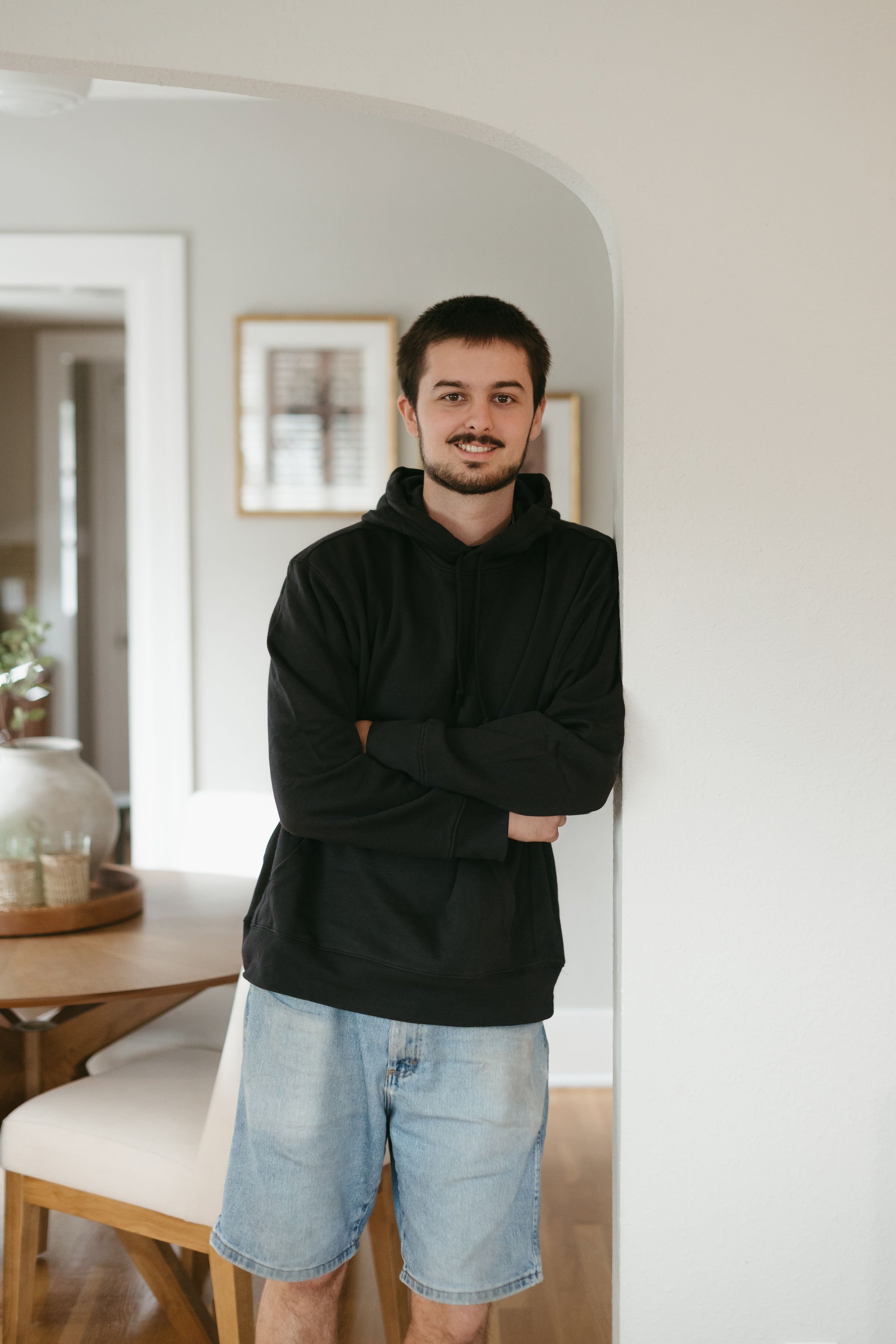 A young man with dark hair and a beard, wearing a black hoodie and light blue denim shorts, stands with arms crossed against a white wall in a living room.