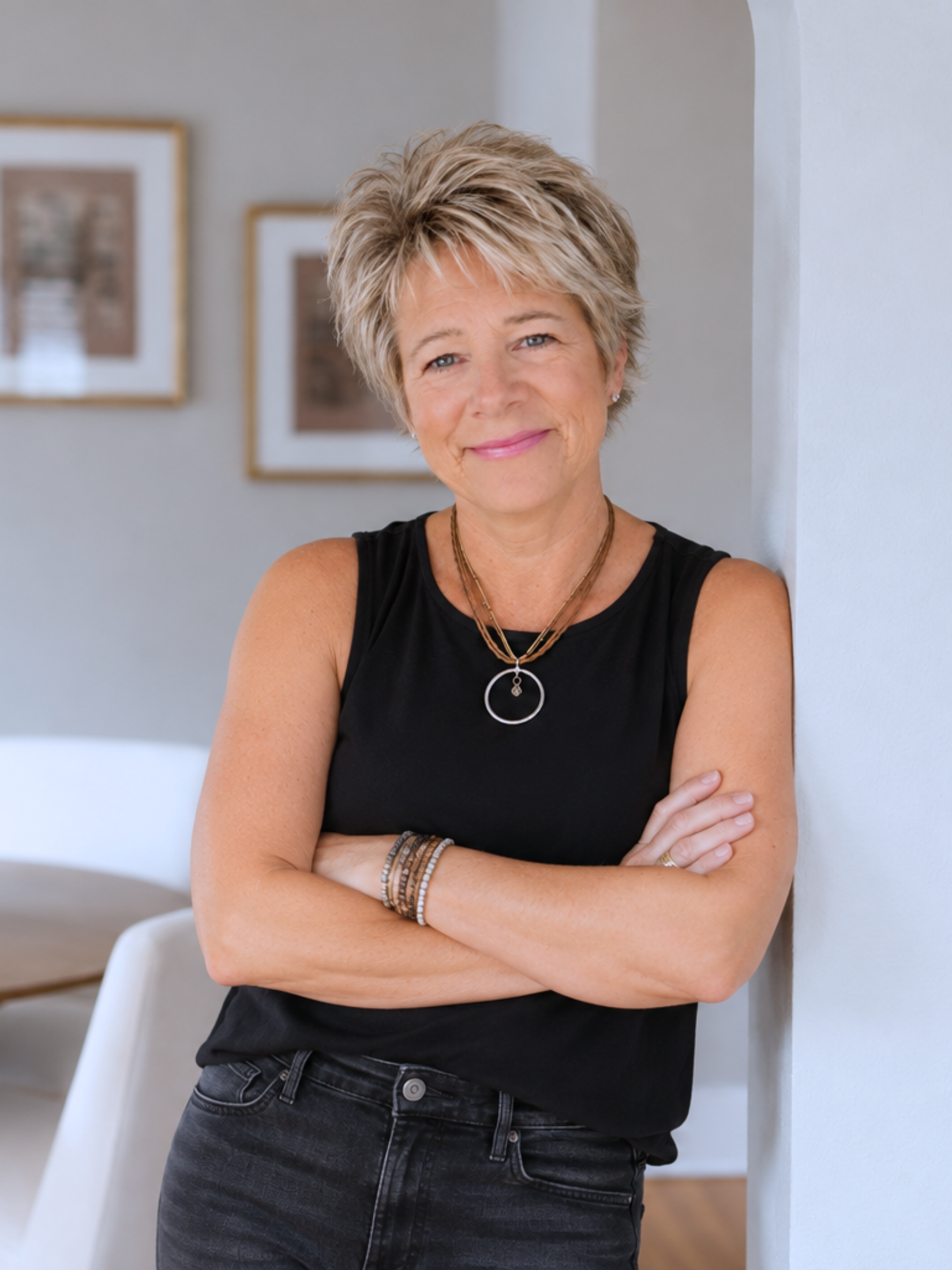 A woman with short, curly blonde hair, wearing a black sleeveless top, Jewelry, and watch, standing with her arms crossed and smiling in a room with framed pictures on the wall.