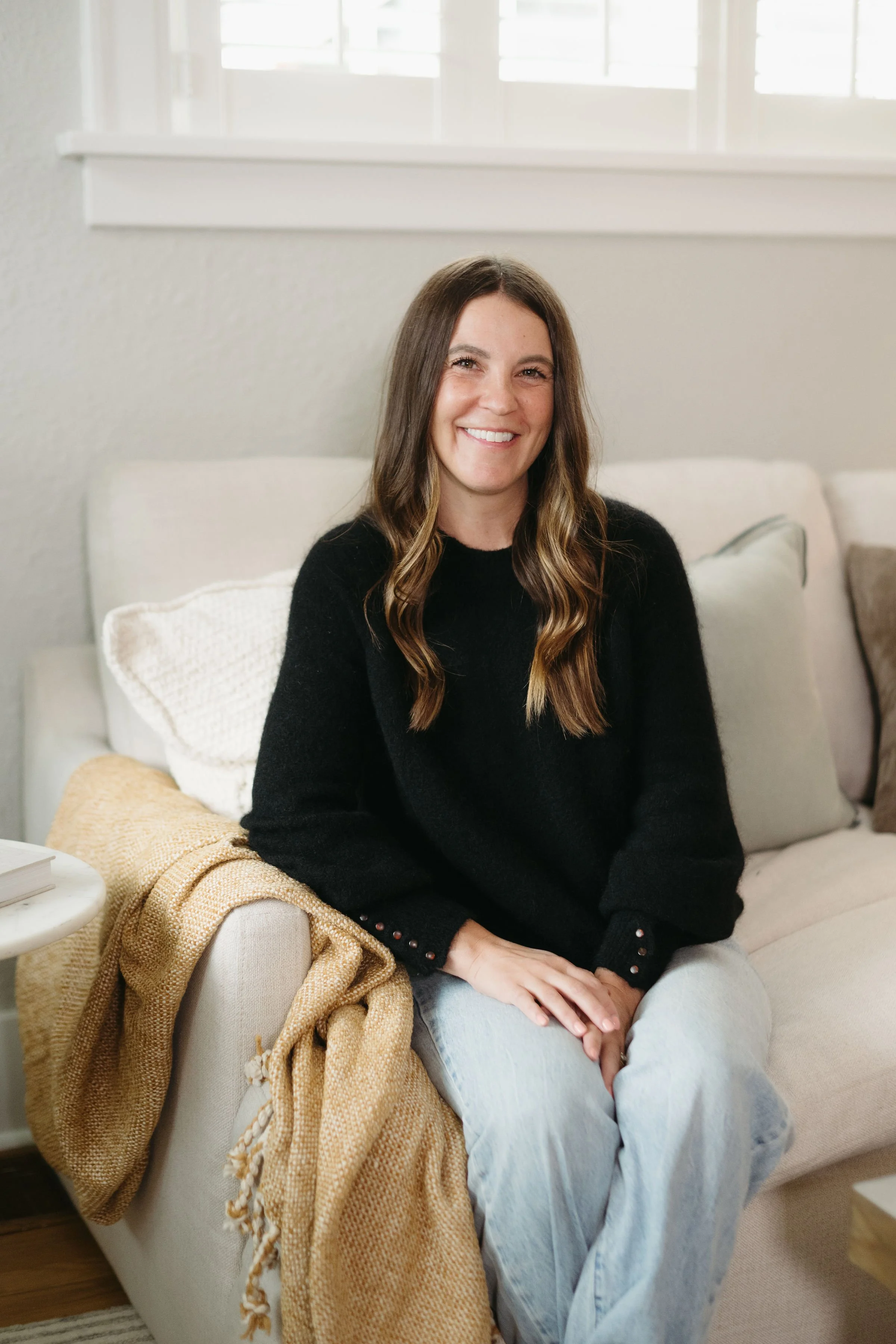 A woman with long brown hair smiling and sitting on a beige sofa, with a yellow blanket draped over the armrest, in a bright room with a window in the background.