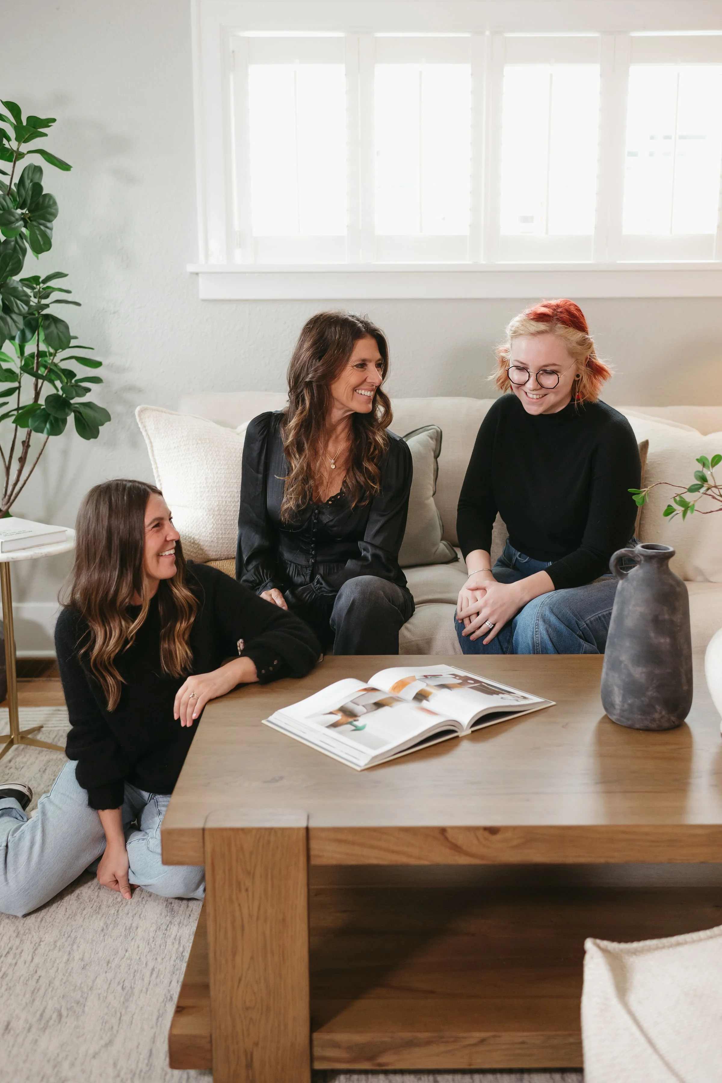 Four women engaging in conversation in a cozy living room with natural light, plants, and a wooden table with an open magazine.