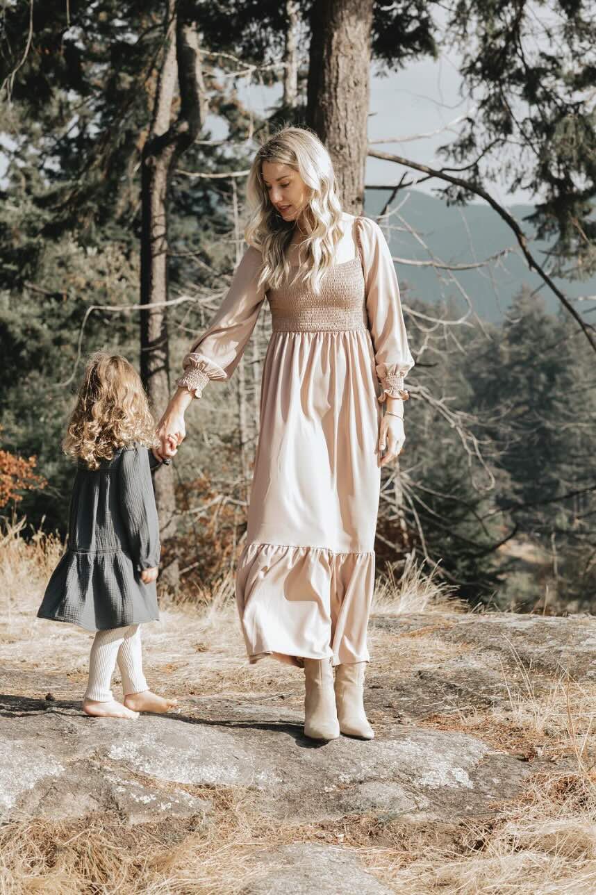 A woman dressed in a long, beige dress holding hands with a young girl in a black dress outside in a wooded area, with trees and mountains in the background.