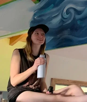 A girl sitting on a surface, holding a white water bottle and a microphone, smiling. She has long hair, is wearing a black cap, and a sleeveless top. There is a colorful ceiling mural behind her with blue swirling patterns.
