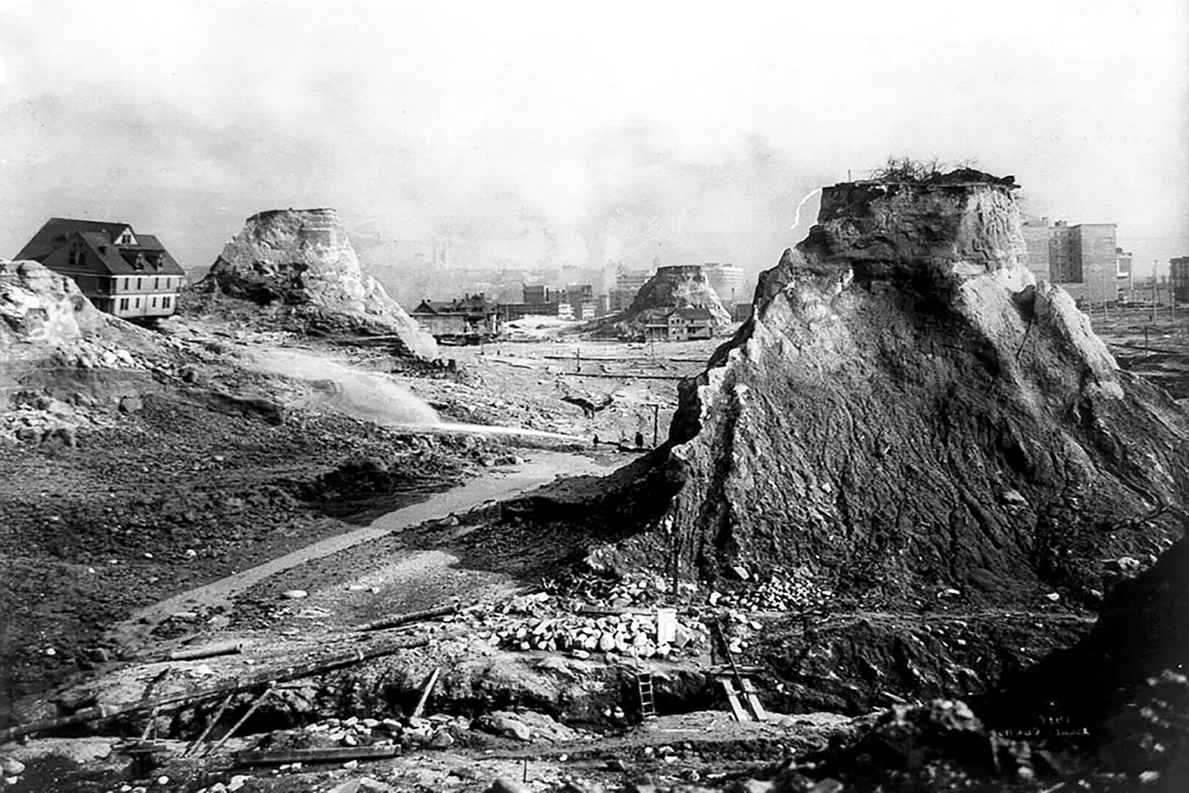 Black and white historic photograph of a construction site with large rocky formations, partially built structures, and dirt roads, possibly from the early 20th century.