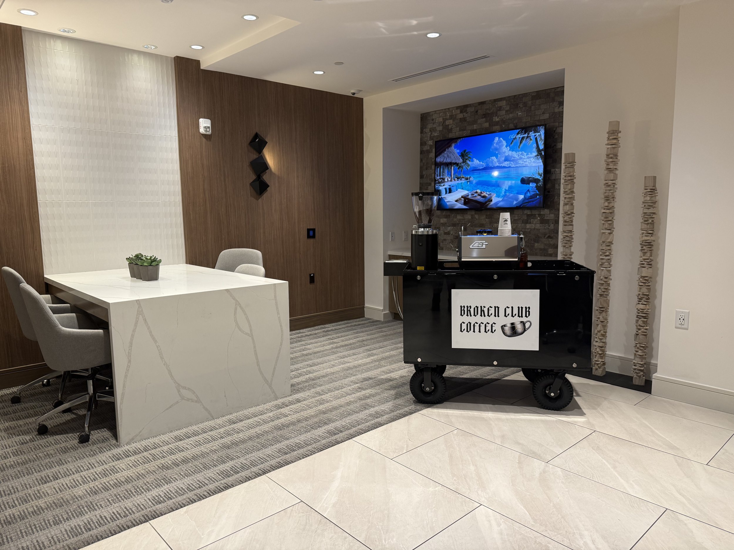 The image shows a modern, stylish indoor lounge area with a dining table, chairs, a large flat-screen TV displaying a beach scene, and decorative elements including a plant, wall art, and wooden sculptures. There is also a sign on a cart that reads "Broken Club Coffee."