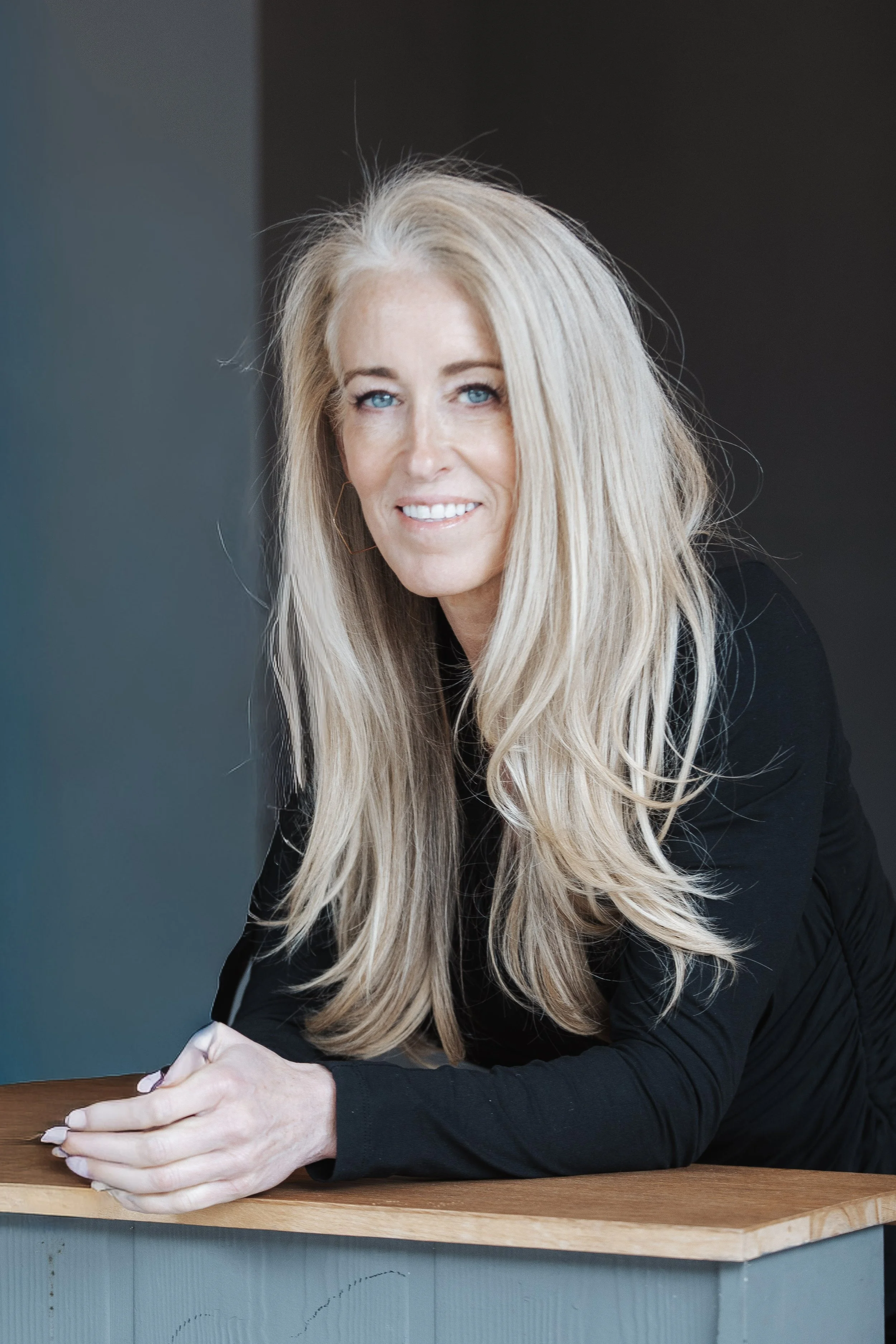 A woman with long blonde hair and blue eyes sitting at a wooden table, smiling slightly, wearing a black top, against a dark and blue background.