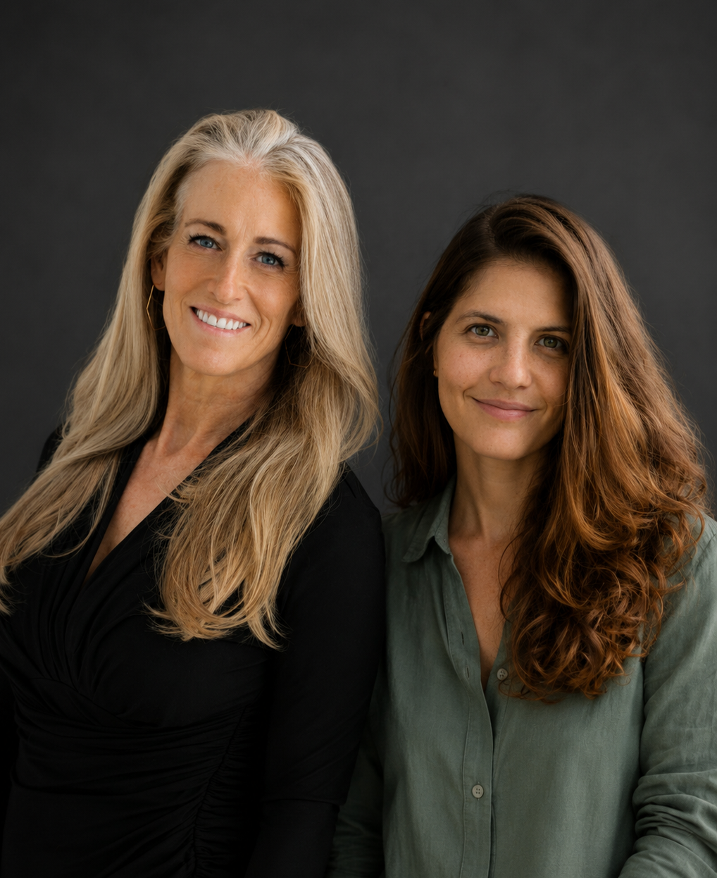 Portrait of two women smiling, one with long blonde hair and the other with long brown hair, against a dark gray background.