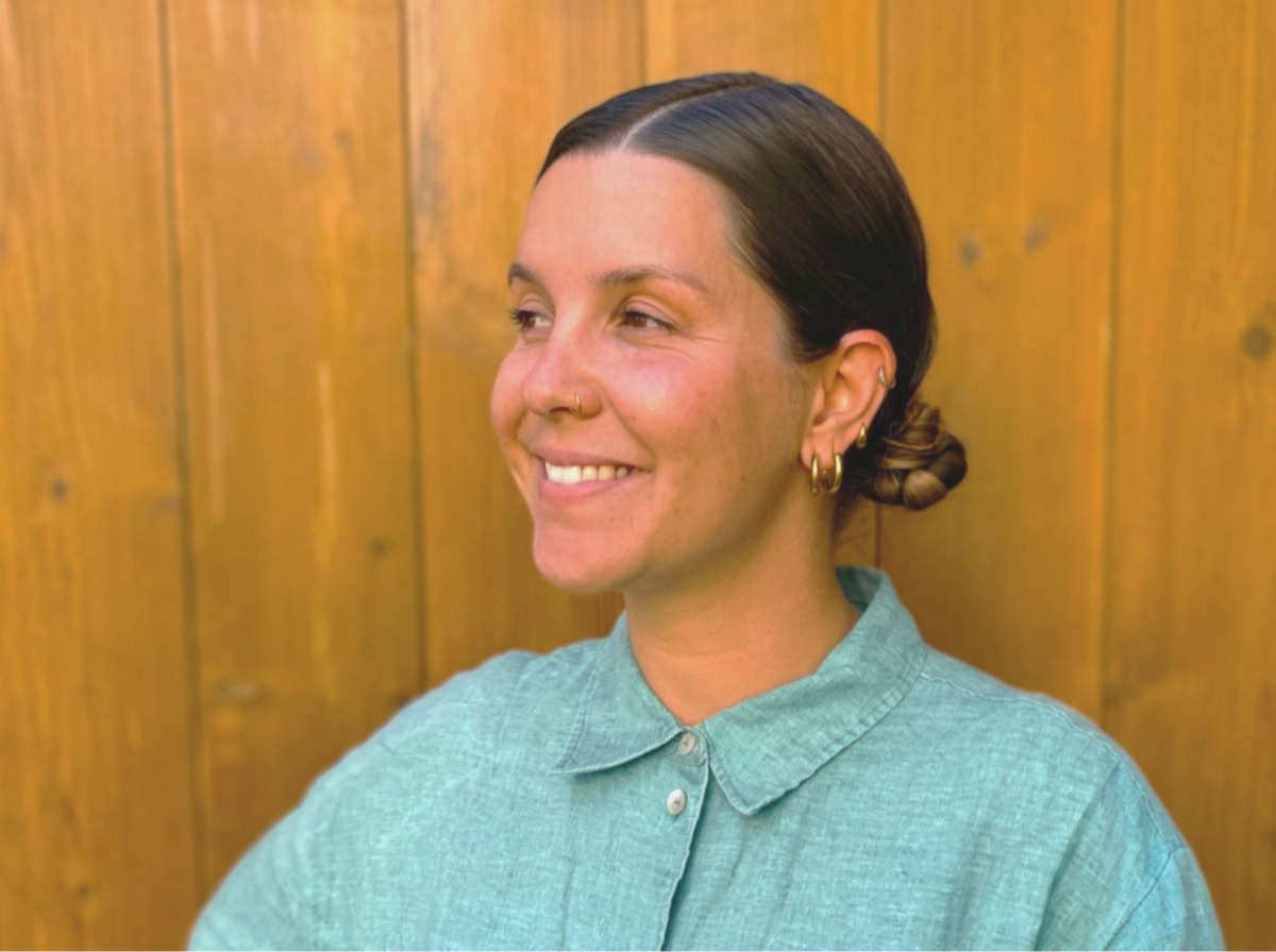 A woman with dark hair styled in a low bun, smiling, wearing a light green button-up shirt, and multiple earrings, standing against a wooden background.