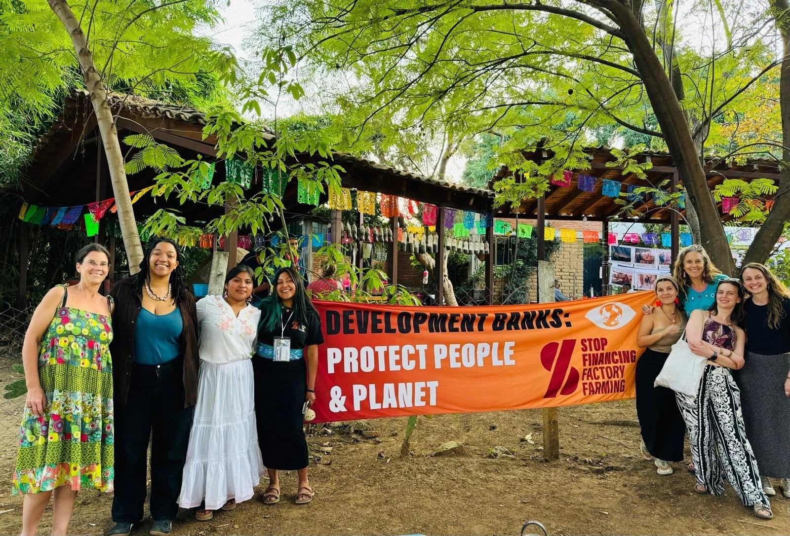 Group of eight women standing outdoors holding a large orange banner with the words "Development Bricks: Protect People & Planet Stop Financing Factory Farming." Behind them are trees, colorful papel picado decorations, and a small structure with photos and decorations.