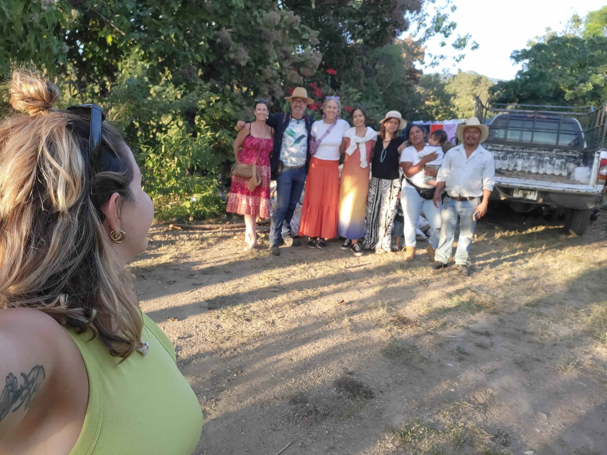 A group of eight people standing outdoors in front of trees and a truck. The group includes men, women, and a child. One woman is taking the photo, and the others are smiling.
