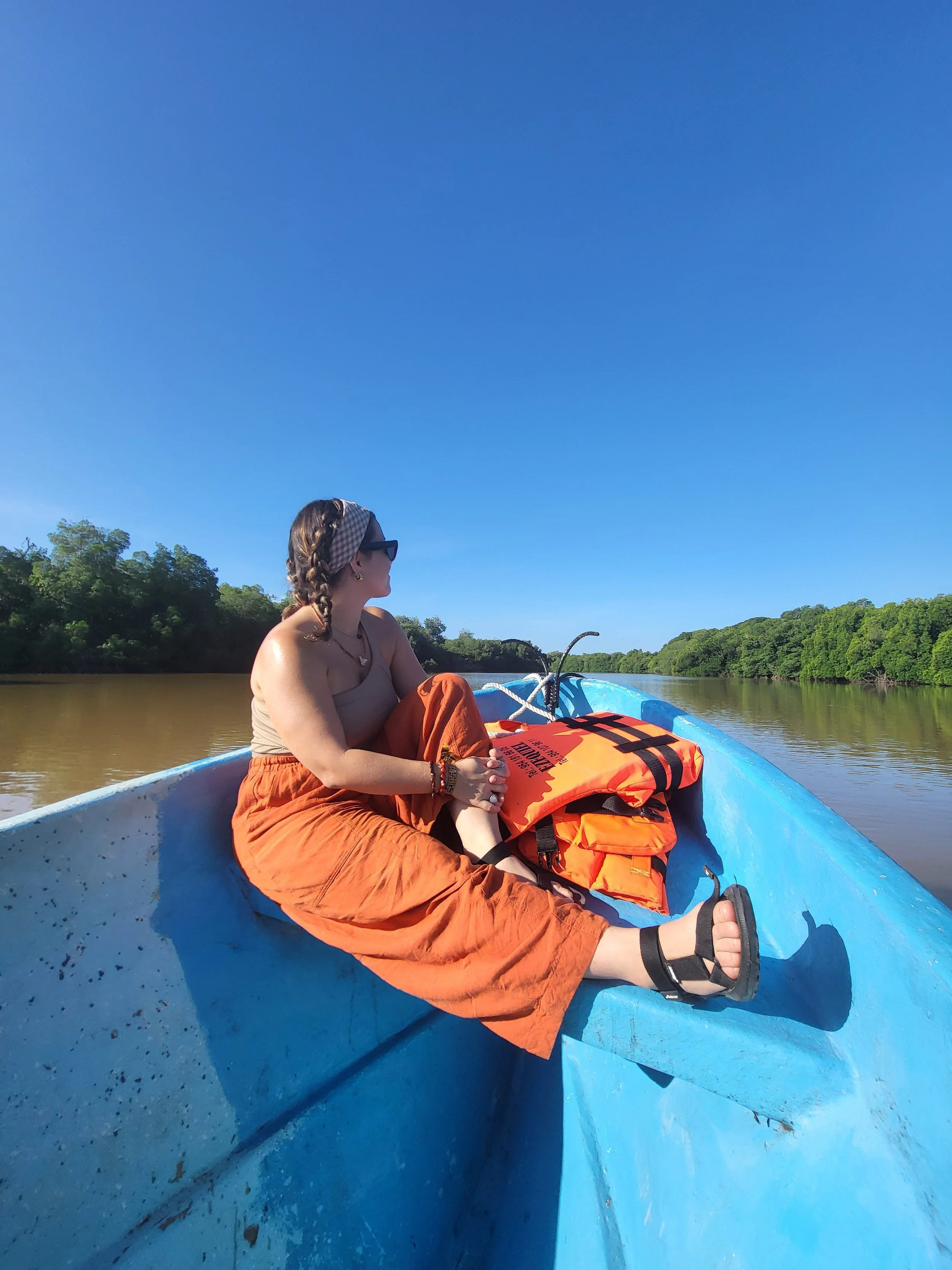 A woman with braided hair, wearing glasses and orange pants, sitting on a blue boat in a river with green trees along the shoreline, under a clear blue sky.
