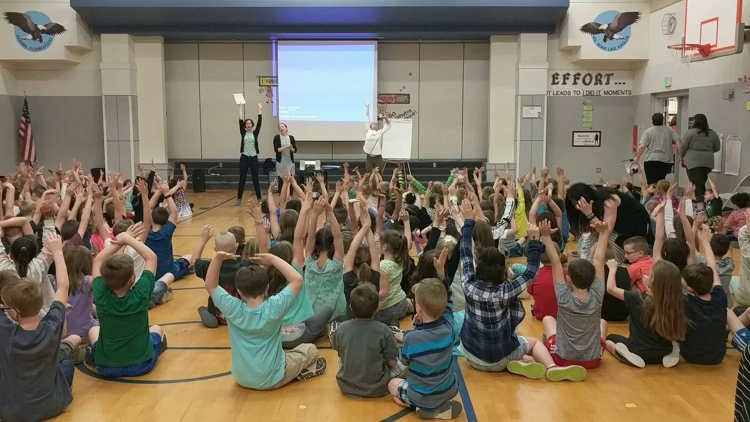 School gymnasium with children sitting on the floor, raising hands, listening to Tracy Subisak on stage in front, some holding papers, with a large projection screen behind them.