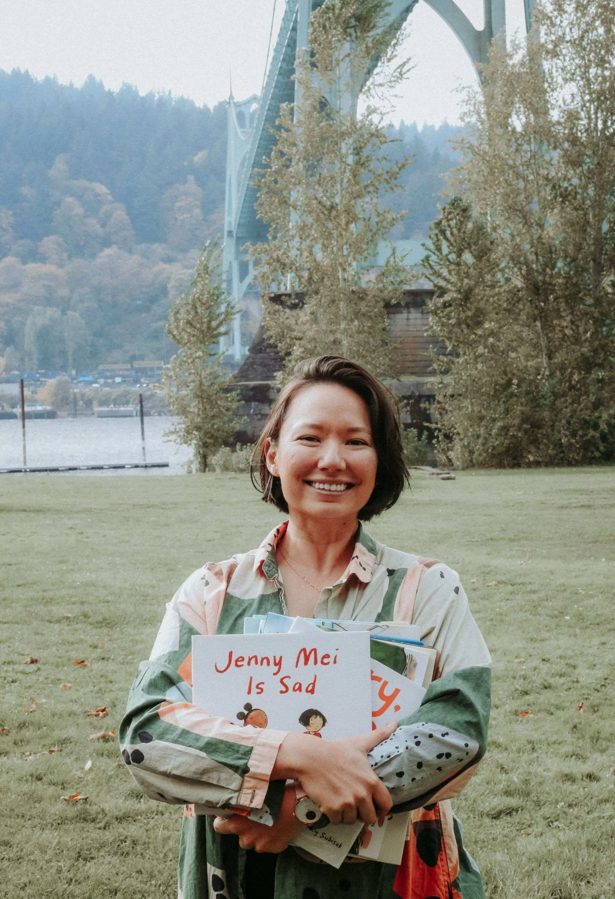 Tracy Subisak smiling and holding books and papers with a sign that reads "Jenny Mei Is Sad." She is standing outdoors on a grassy area with trees and a bridge in the background, under an overcast sky.