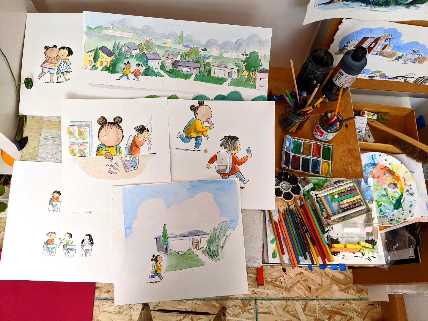 A workspace with several watercolor and ink illustrations of children, scenes of kids playing, and outdoor settings. Art supplies including watercolor paints, colored pencils, brushes, and jars are scattered on the desk.