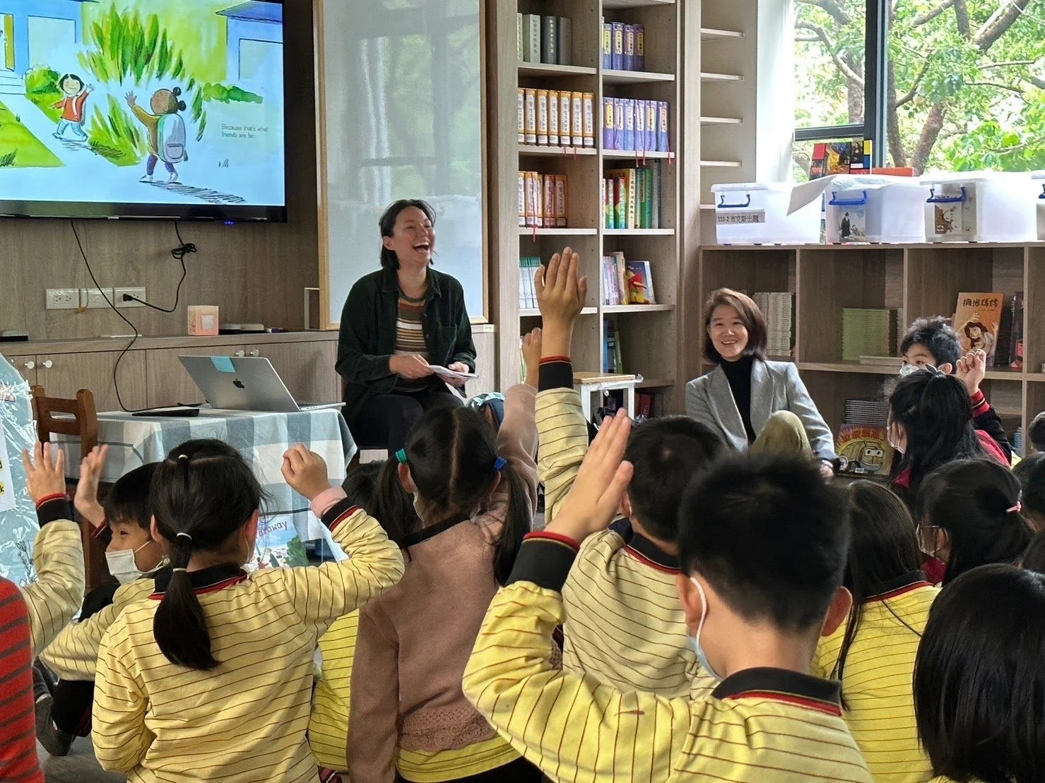 A classroom with children wearing yellow and red striped uniforms, some raising their hands.  sits at the front with a laptop, engaging with the students. Several children and a woman in a gray blazer are smiling and listening. The room has bookshelves and a window showing green trees outside.