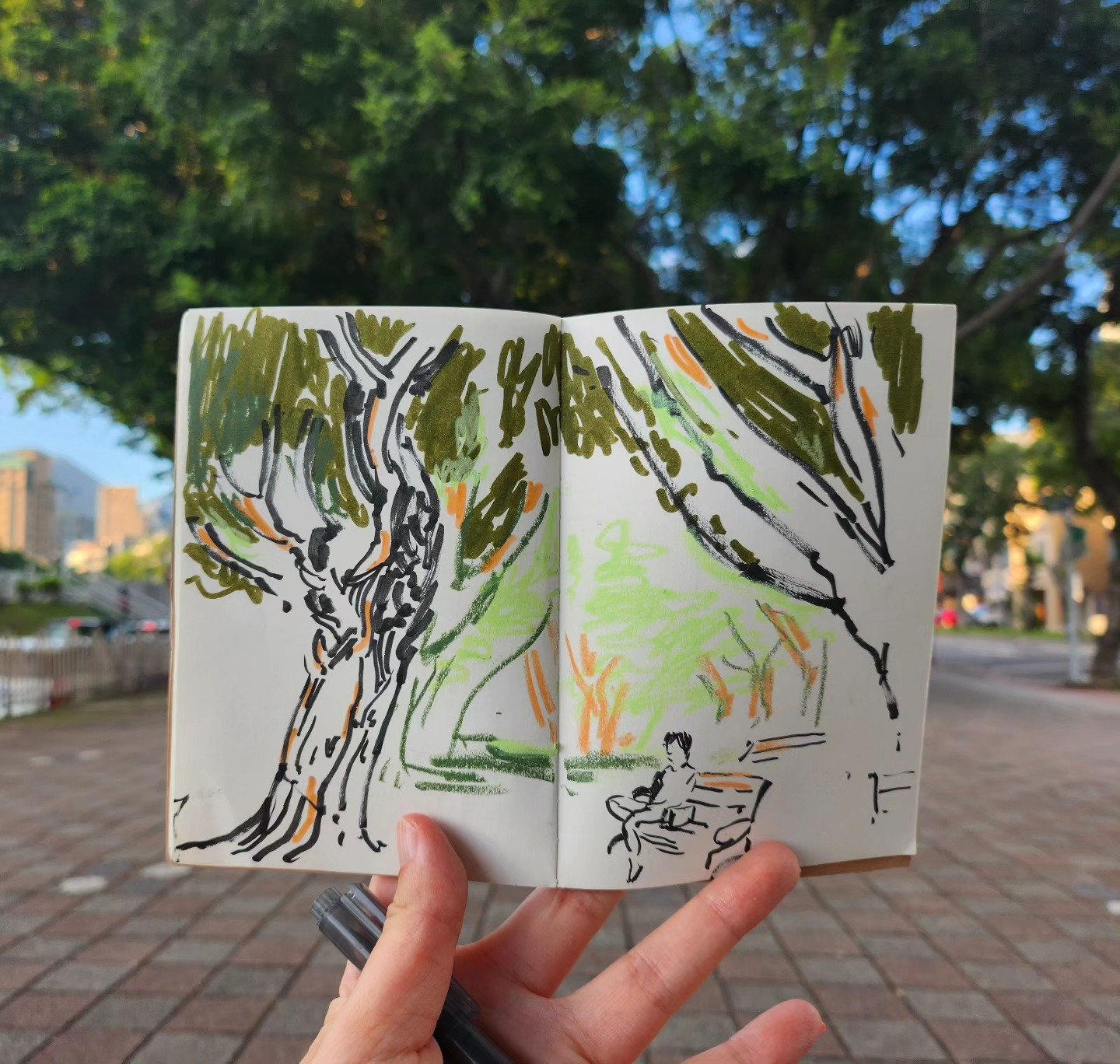 A sketchbook showing a drawing of trees, grass, and a person sitting on a bench, with a background of a park and trees.