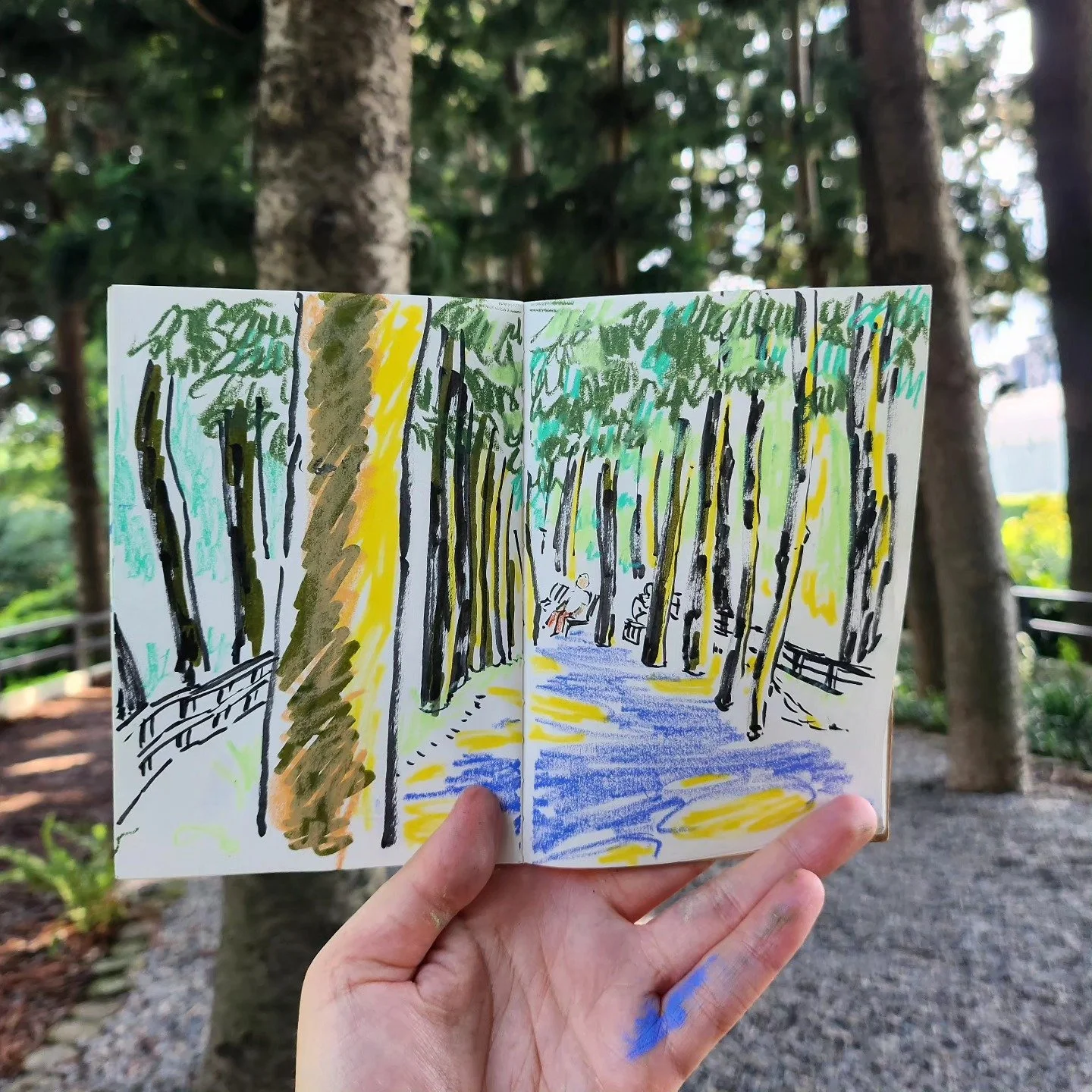 A hand holding a sketchbook with a colorful drawing of a forest path, with tall trees and two people sitting on a bench, in a wooded area.