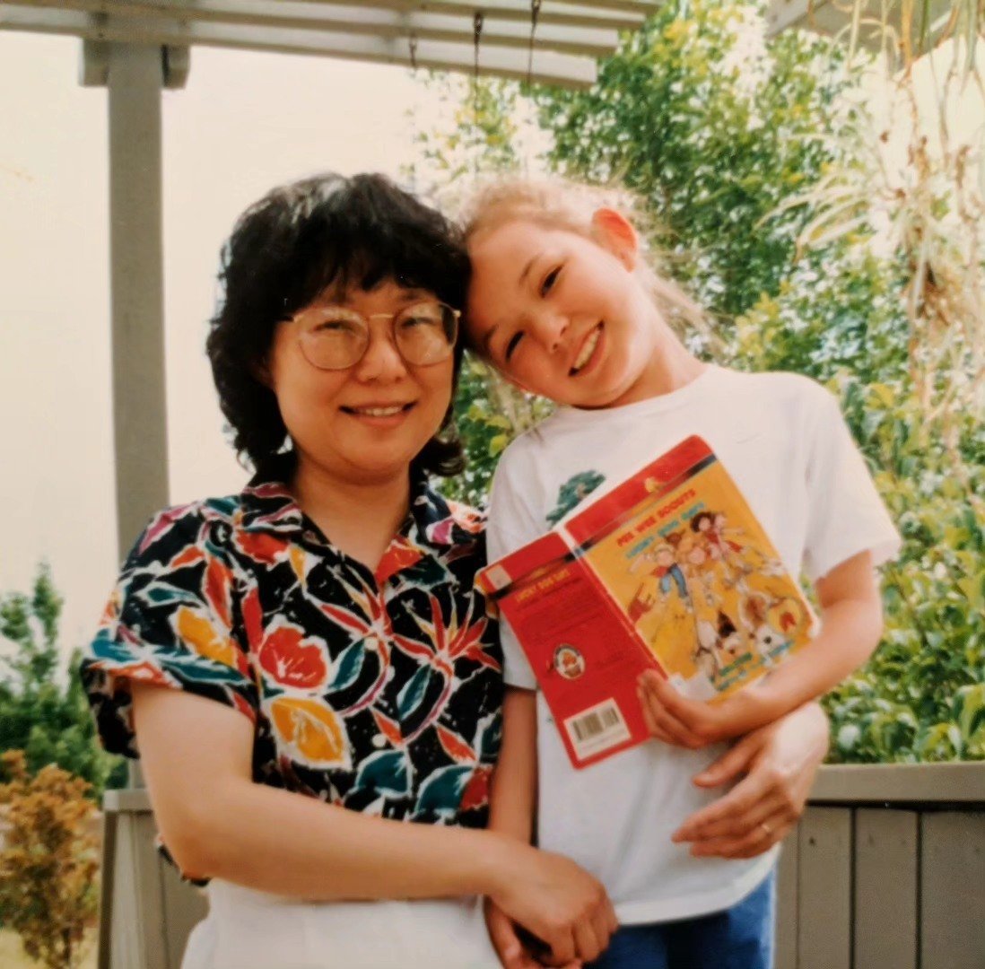 Tracy Subisak as a little girl smiling and hugging her mom outdoors, while holding a book with colorful illustrations.