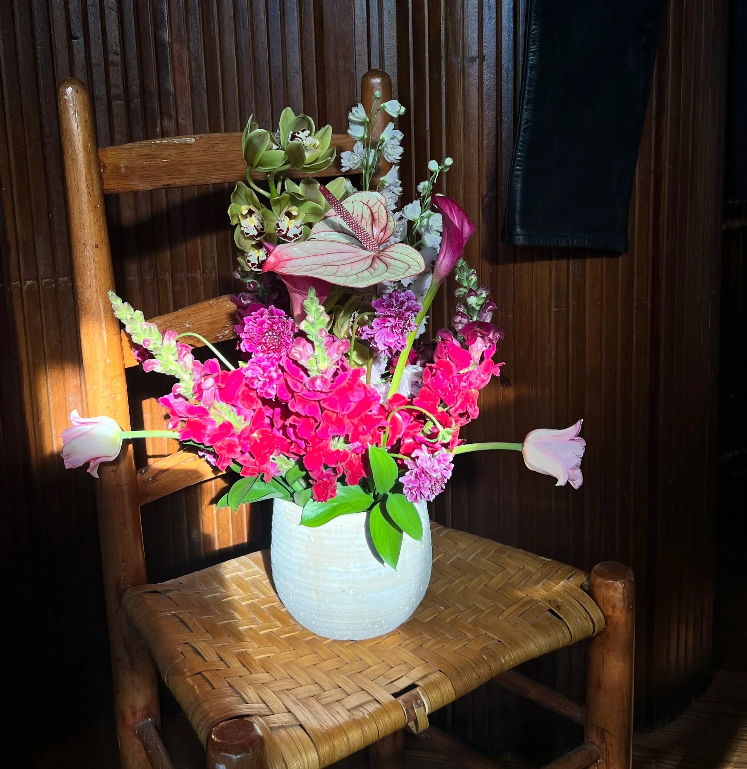 A bouquet of colorful flowers in a white ceramic vase placed on a wooden chair against a wood-paneled wall.