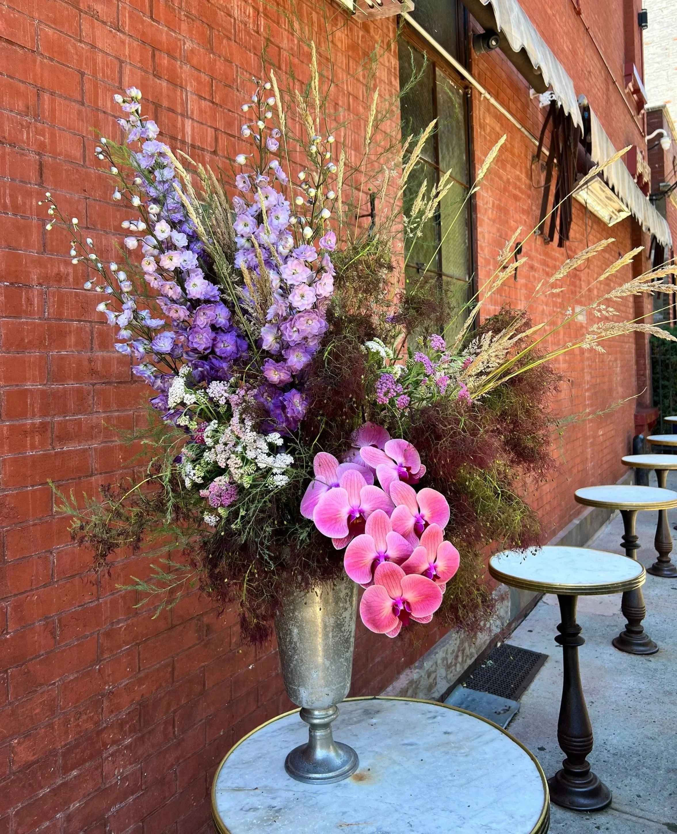 A tall metallic vase filled with purple and pink flowers is placed on a round white marble-topped table outside a brick building. Several chairs are visible along the sidewalk, and an outdoor awning with striped fabric extends above the window.