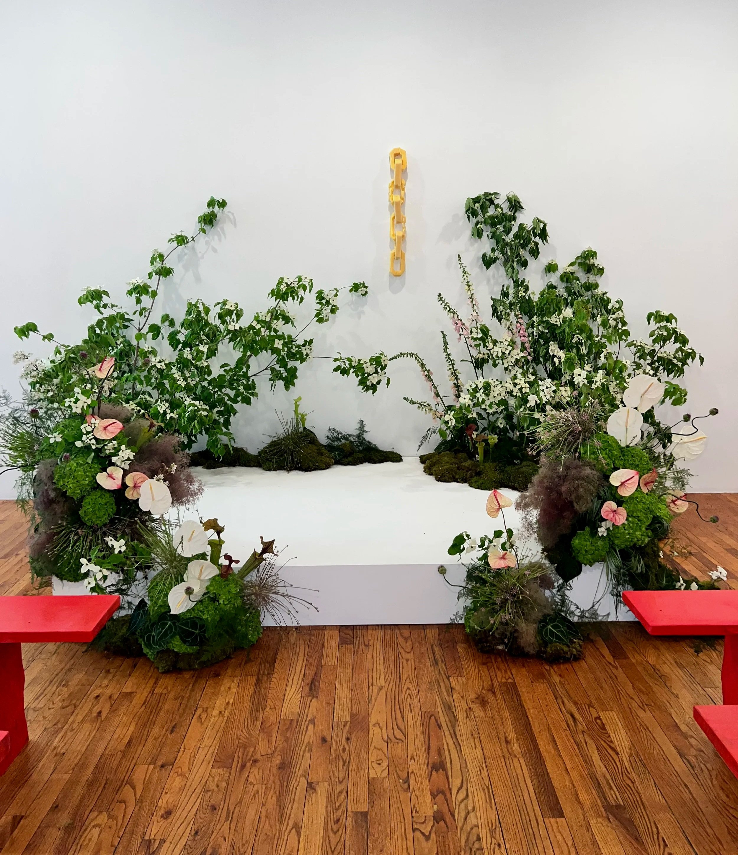 A white platform with various green plants and flowers arranged around it, with red benches on either side, and a plain white wall with a yellow abstract art piece hanging in the center.
