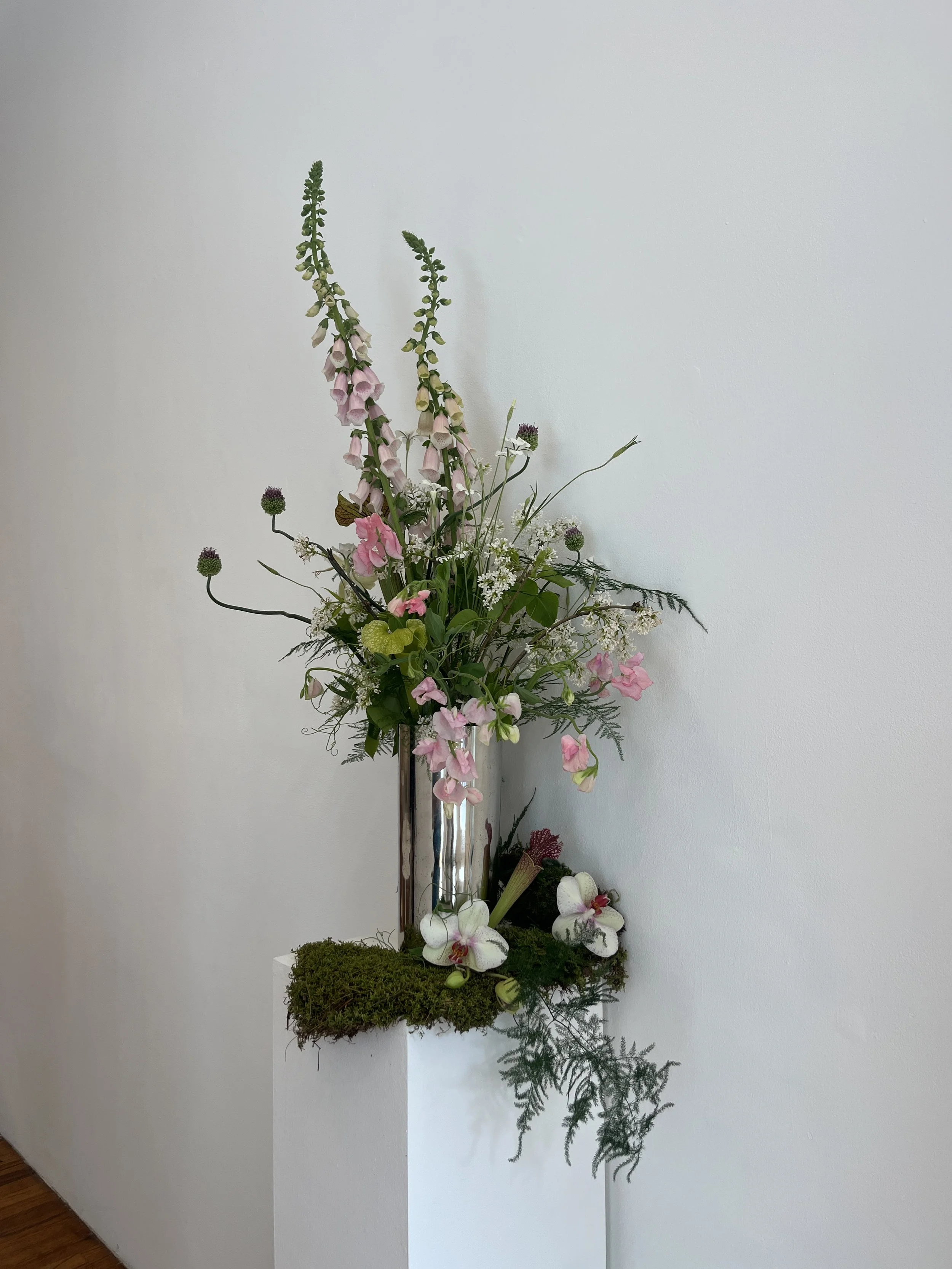 A floral arrangement in a tall glass vase with pink and white flowers, green foliage, and moss at the base, positioned against a plain white wall.